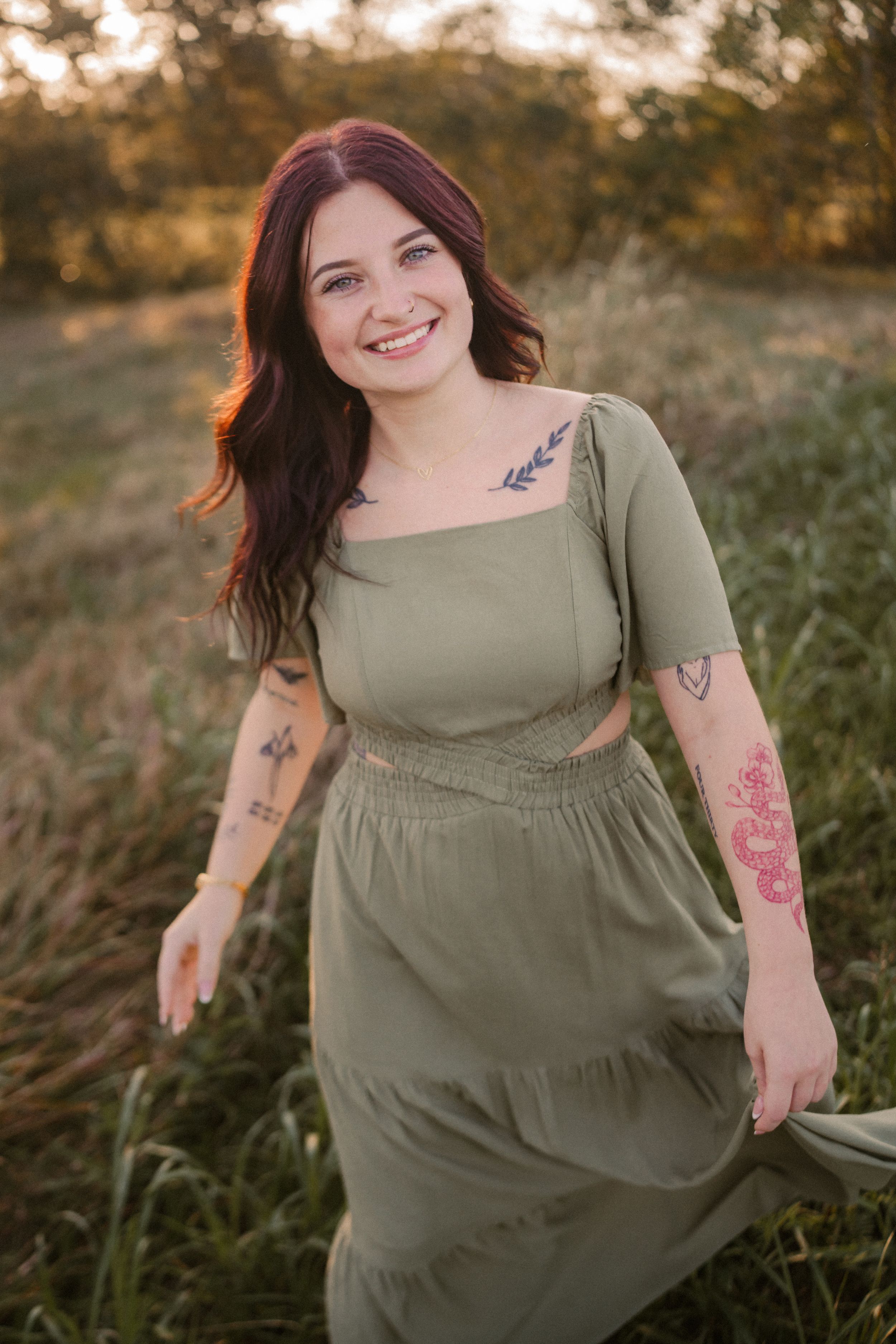 A person in a sage green dress with visible tattoos poses outdoors during golden hour in a grassy field.
