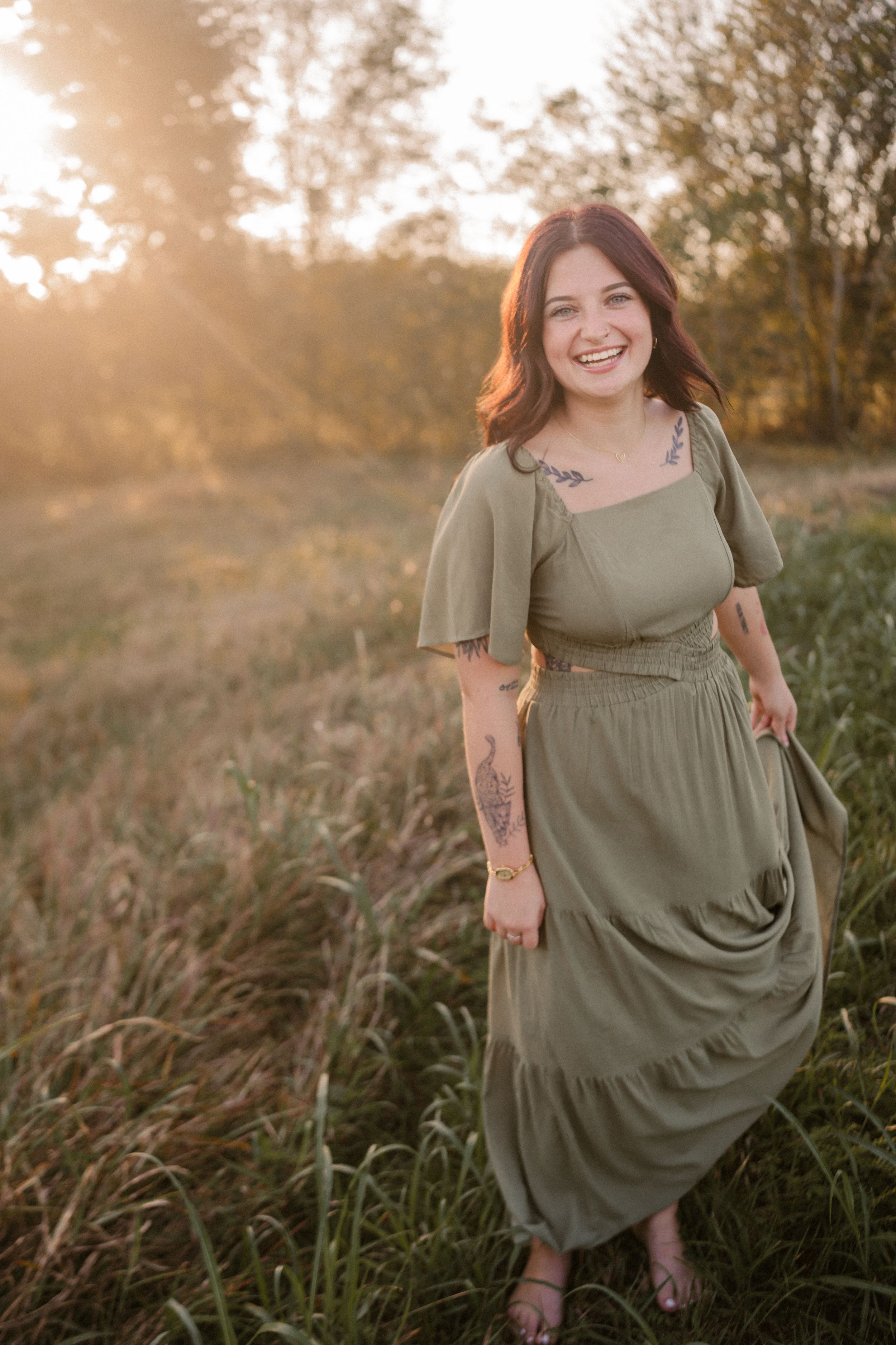 A person in a flowing sage green dress stands in a sunlit field at golden hour with tall grass around them.