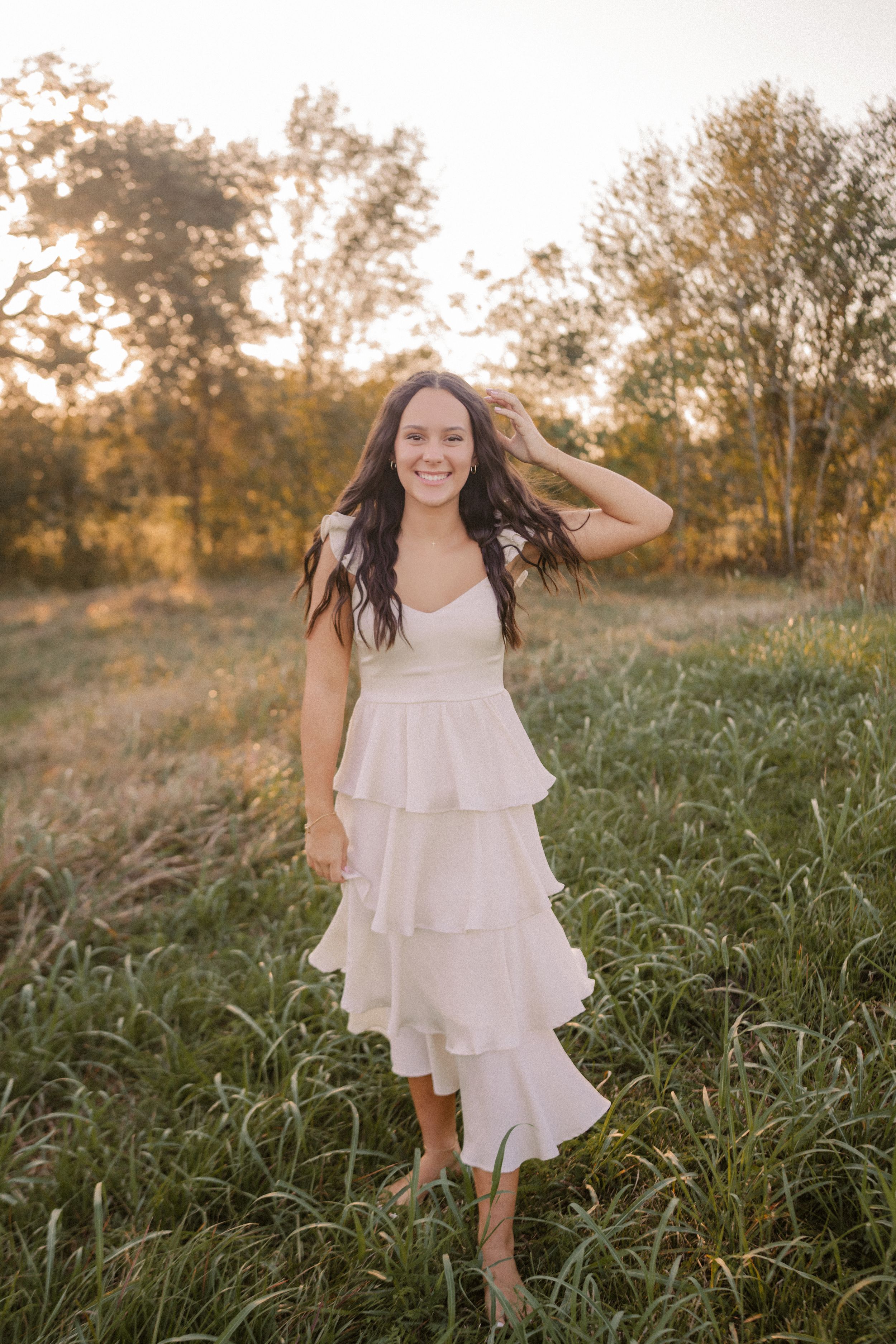 A person wearing a white tiered ruffle dress stands in a grassy field at sunset with golden light filtering through trees.