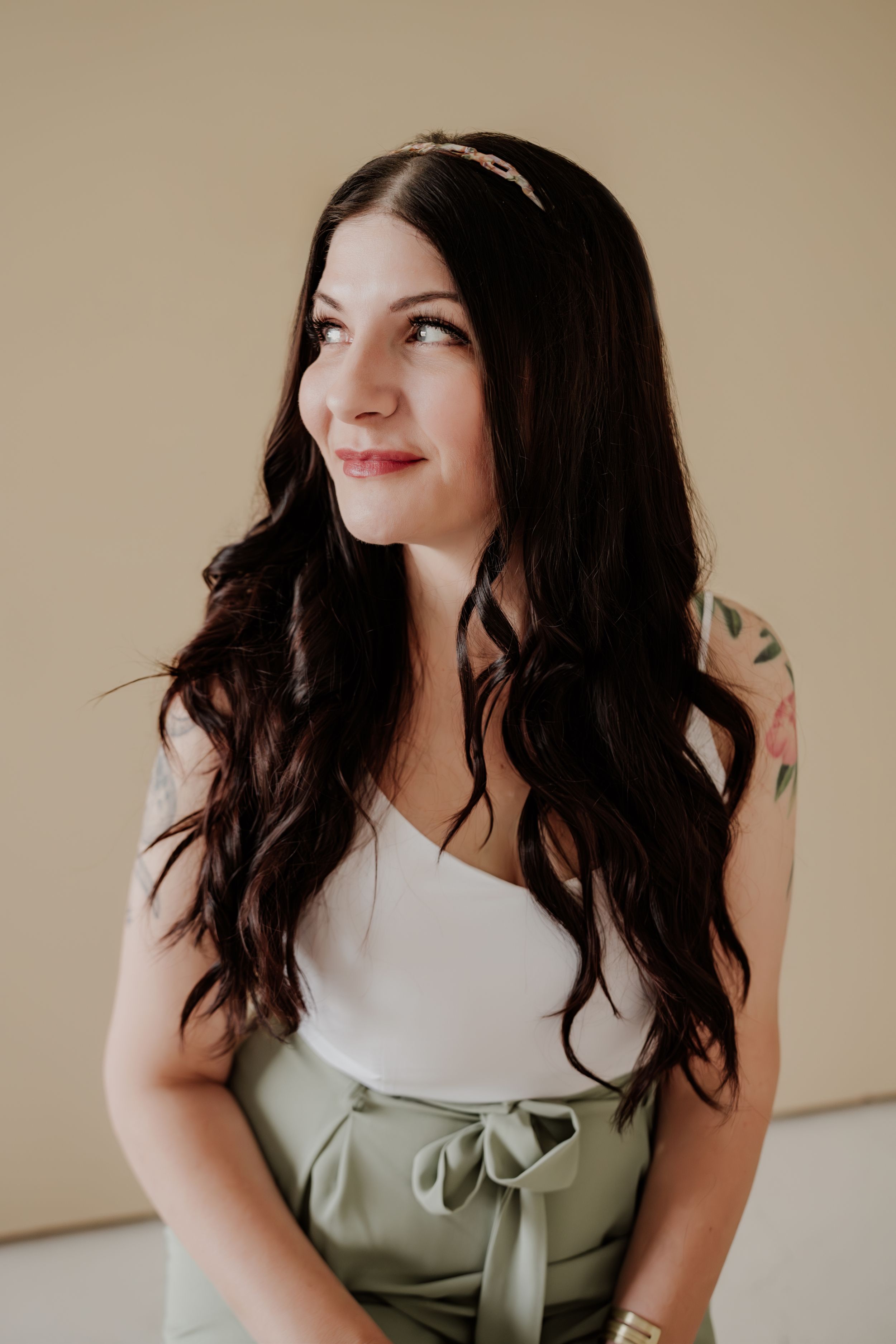 Young woman with long dark wavy hair wearing a white top and sage green skirt looks to the side with a soft smile.