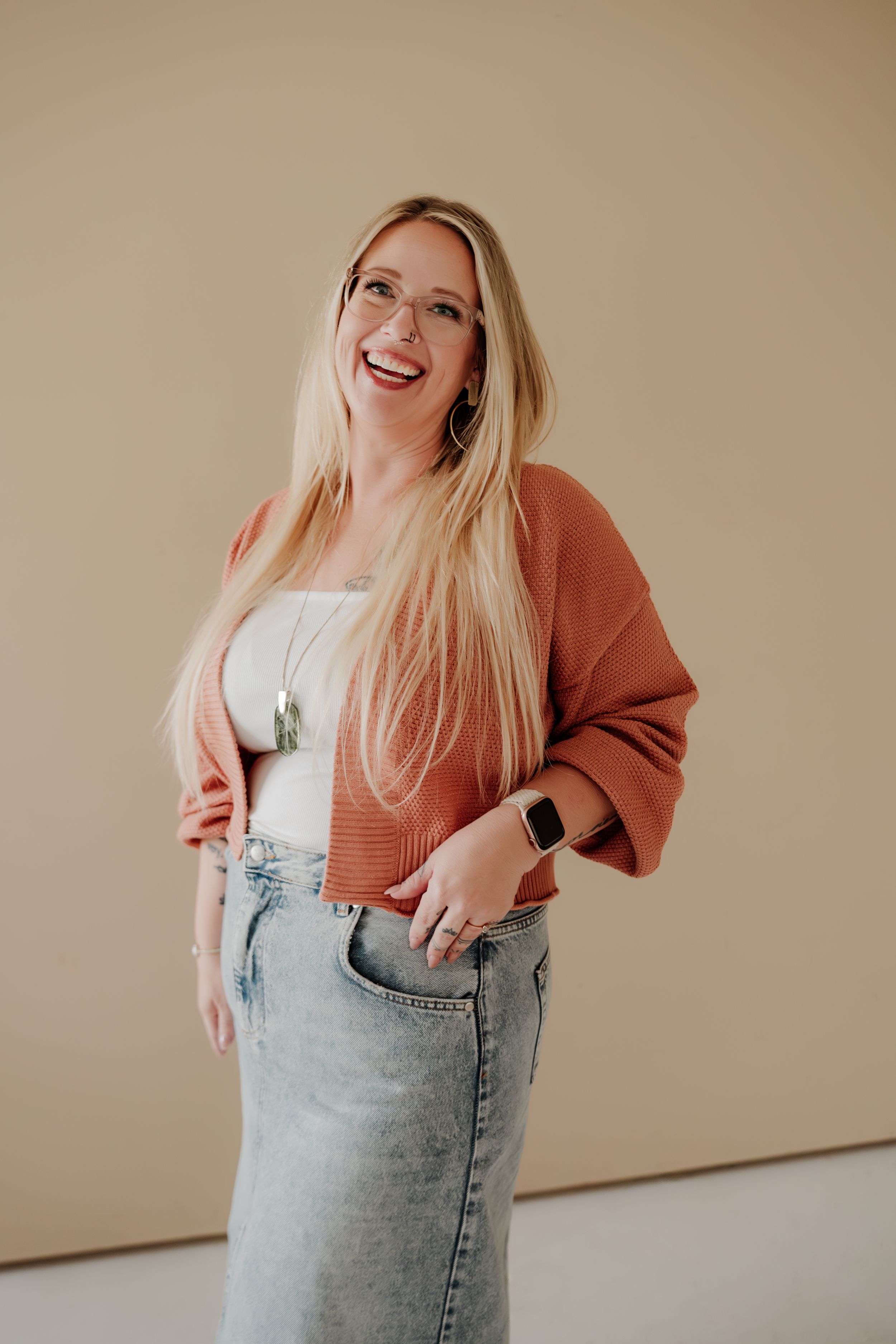 A woman wearing a rust-colored cardigan and jeans poses against a beige wall with a cheerful expression.