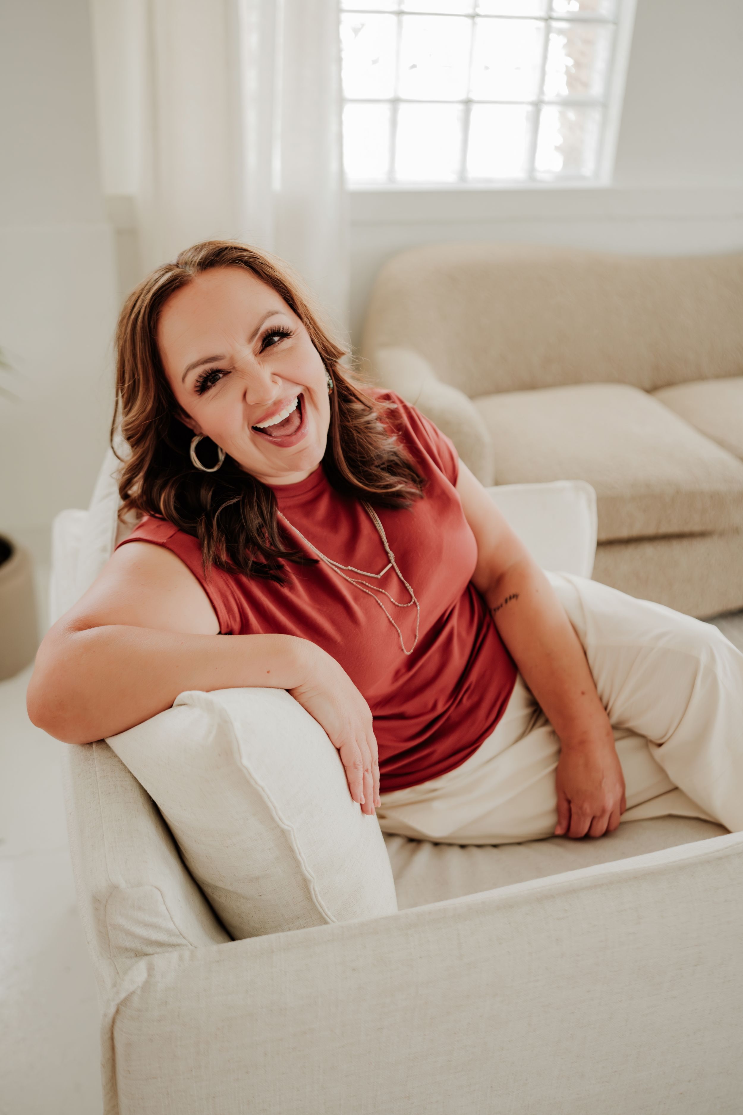 A joyful woman in a red sleeveless top relaxes on a beige couch in a bright, sunlit living room.