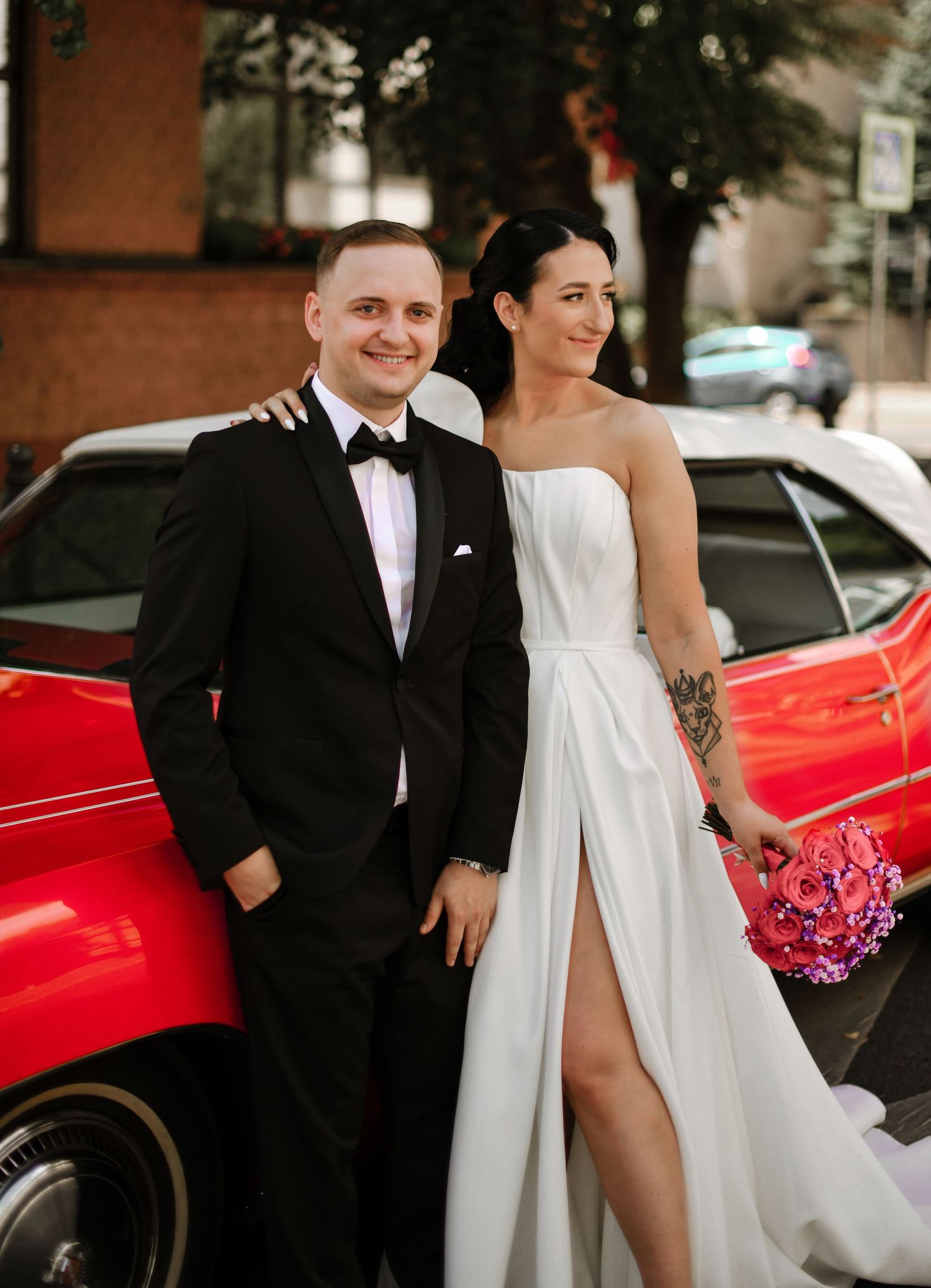 Wedding couple posing beside a vintage red car in urban setting.