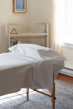 A massage table covered in crisp white linens sits in a peaceful treatment room with natural lighting.