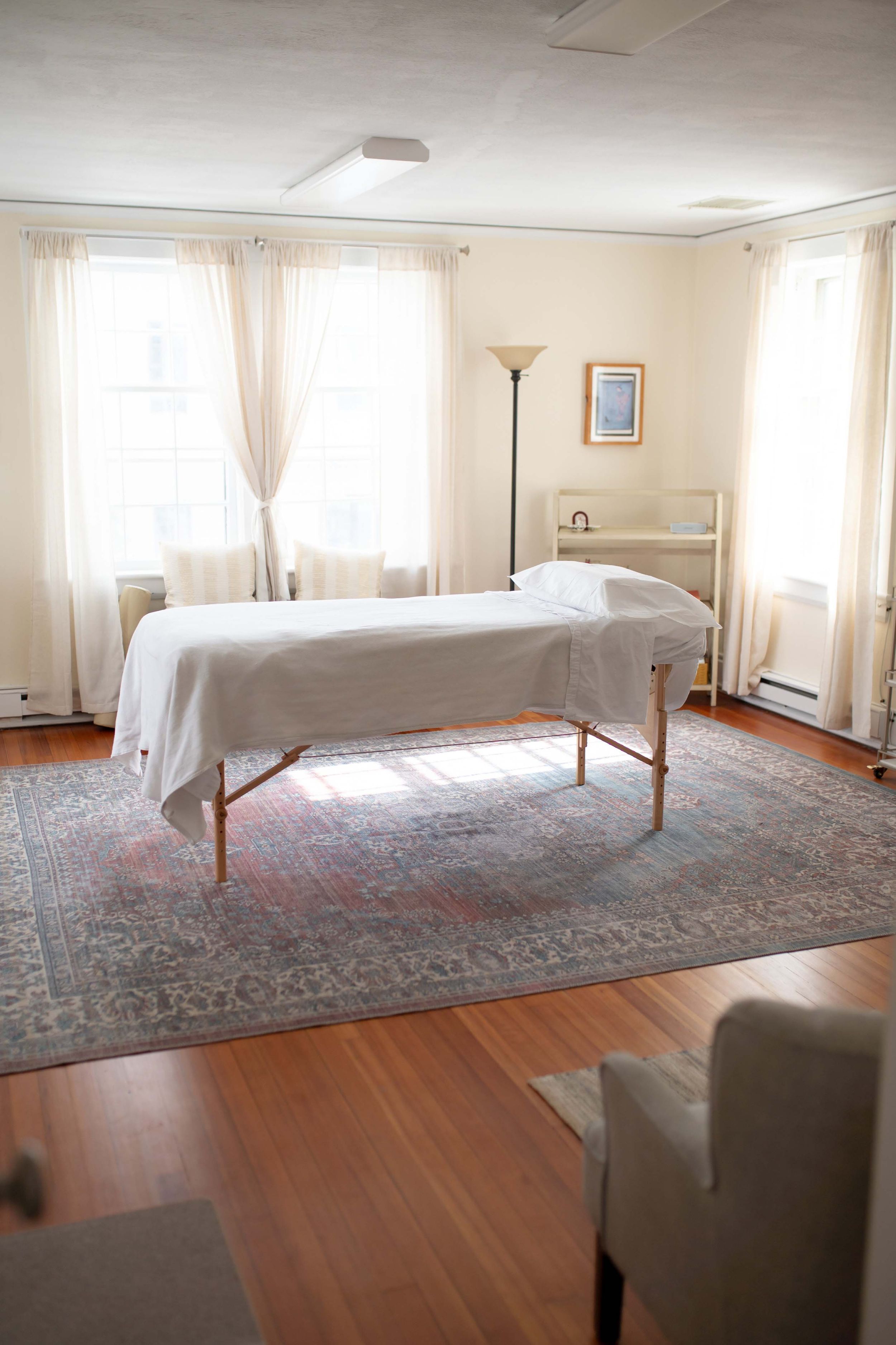 A serene massage therapy room with a treatment table, oriental rug, and natural light streaming through white curtained windows.