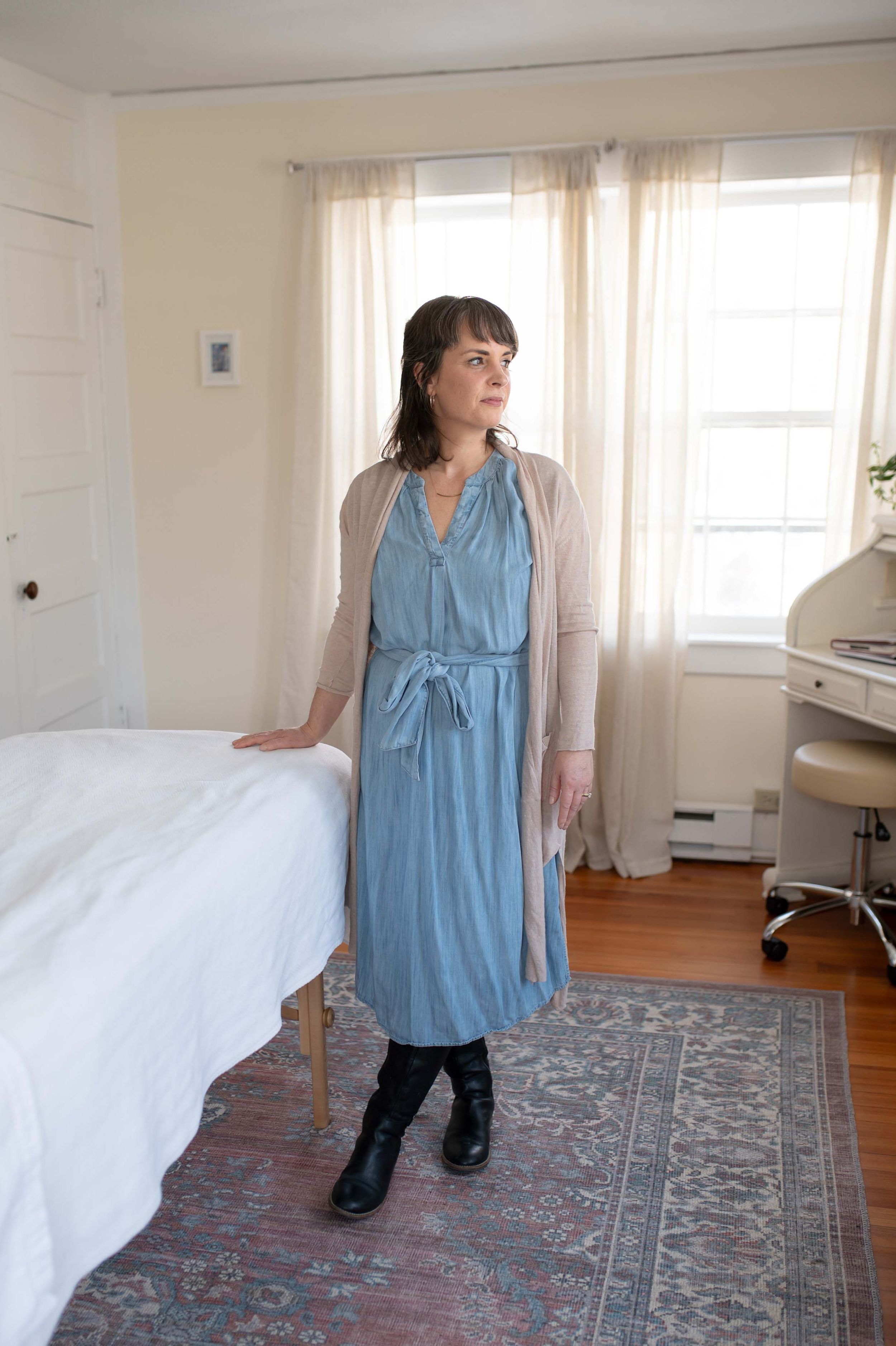 Standing figure in a blue belted dress and beige cardigan sweater in a bright bedroom with white curtains and ornate rug.