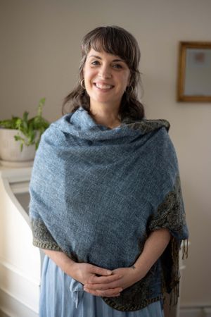 Person wearing a blue knit shawl with dark trim stands smiling in a bright indoor space with houseplants.