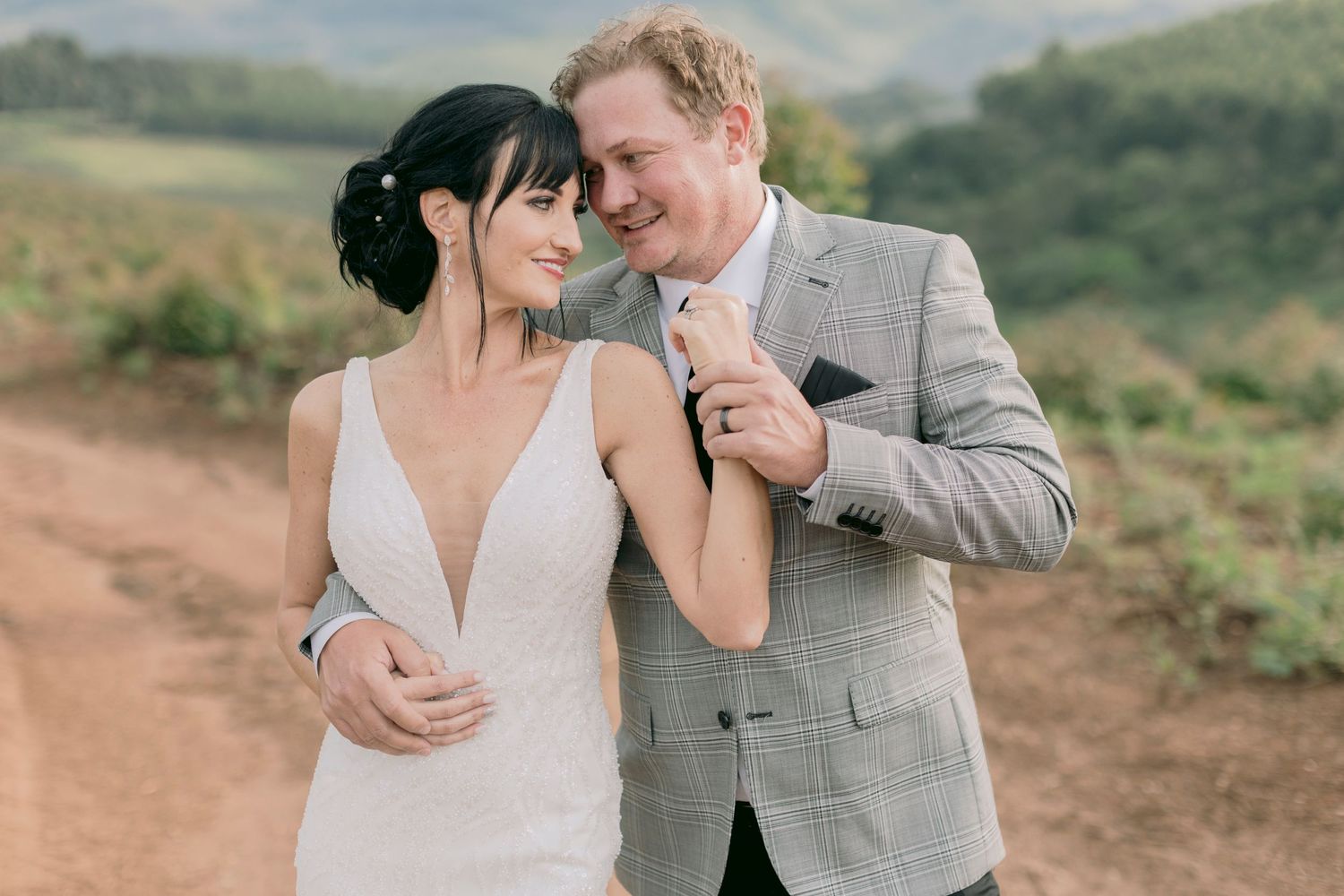 Happy bride and groom walking on a dirt road with scenic green hills in the background.