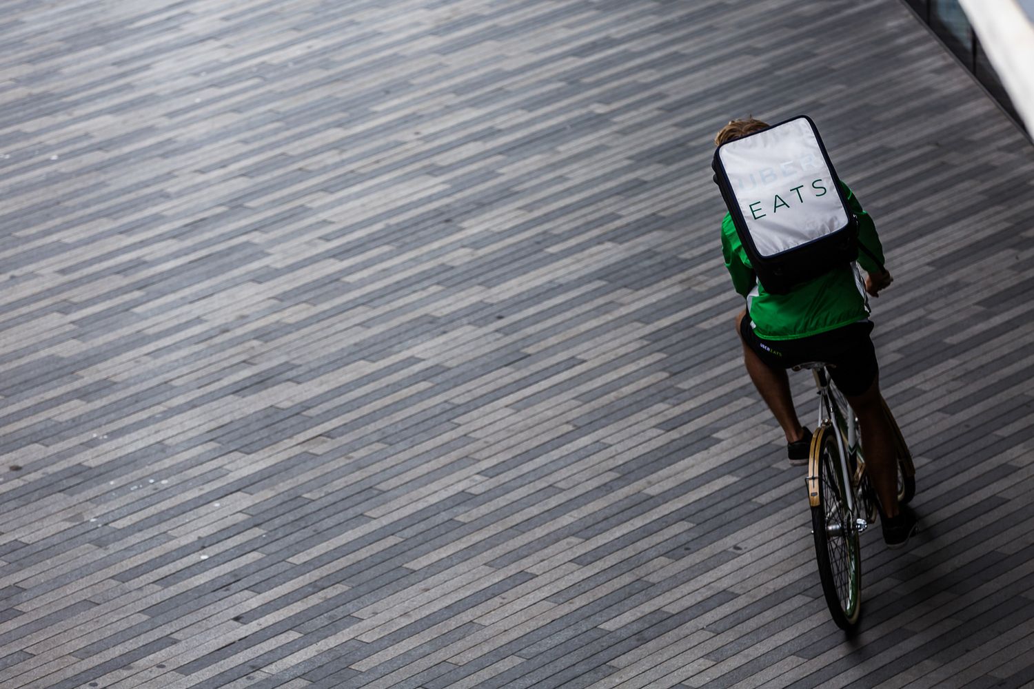 A delivery cyclist with a branded green backpack rides on a textured gray surface from an aerial view.