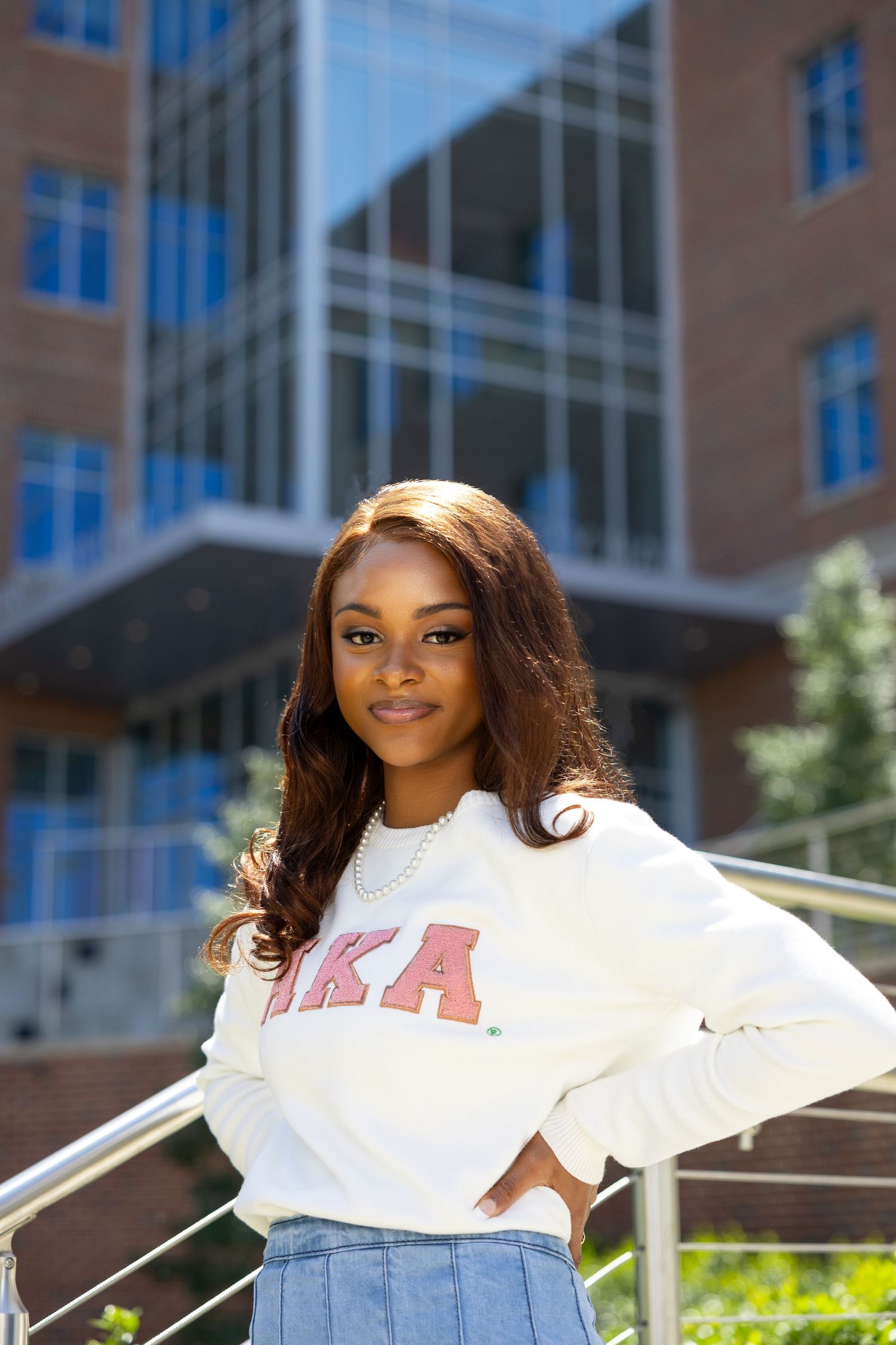 A student wearing a white AKA sweatshirt and denim skirt poses outdoors near a modern campus building.