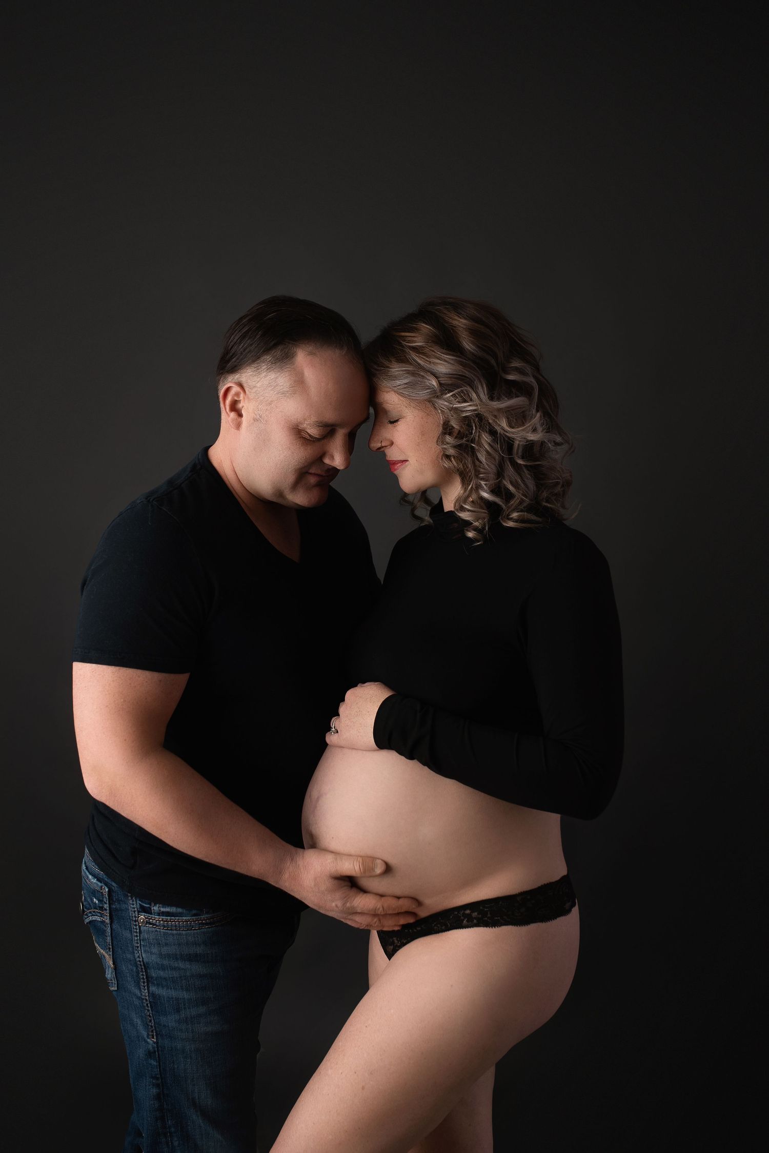 Intimate maternity photo shows couple in black clothing embracing against dark background with tender moment.