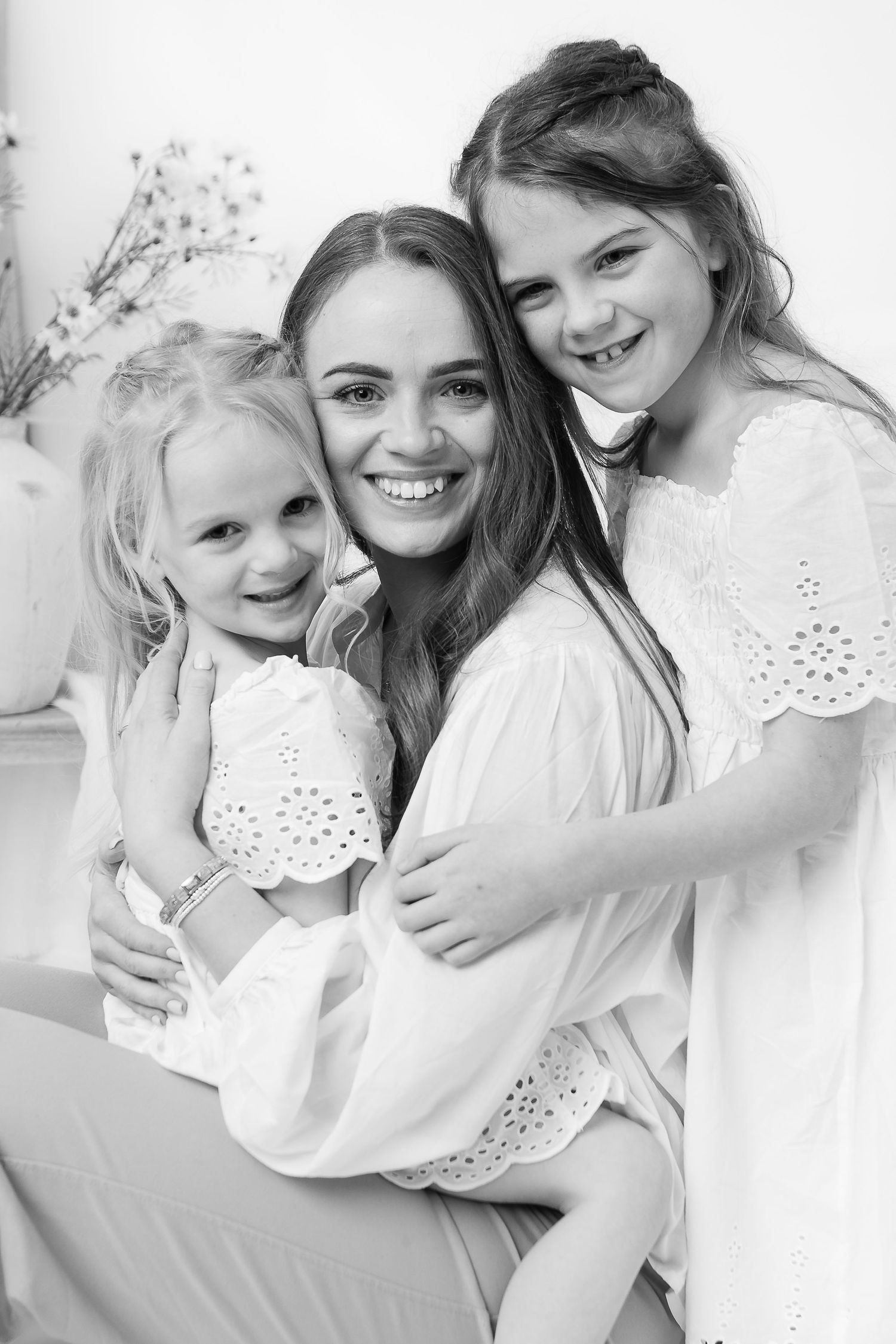 Black and white portrait of three joyful subjects wearing white eyelet dresses while embracing and smiling.