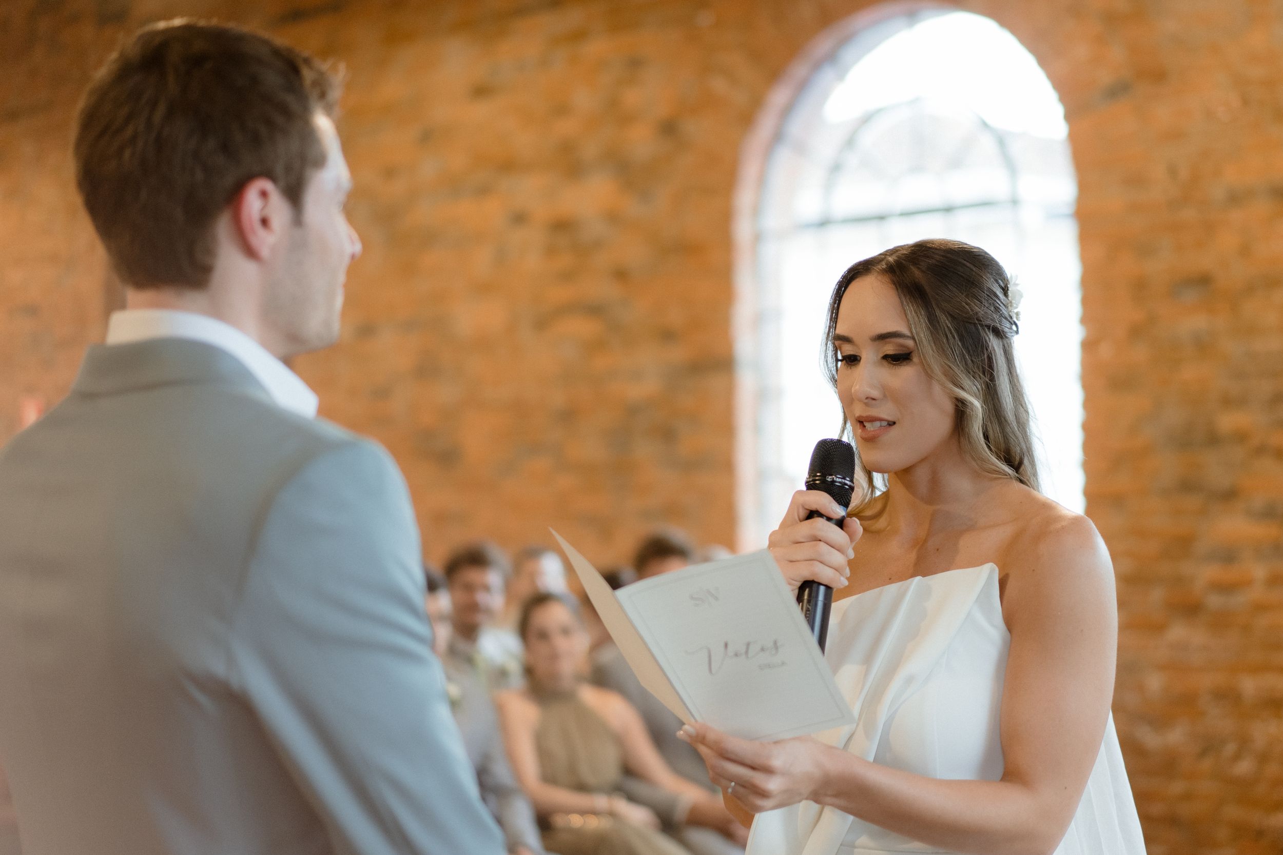 Wedding ceremony with couple exchanging vows in front of exposed brick wall and arched window.
