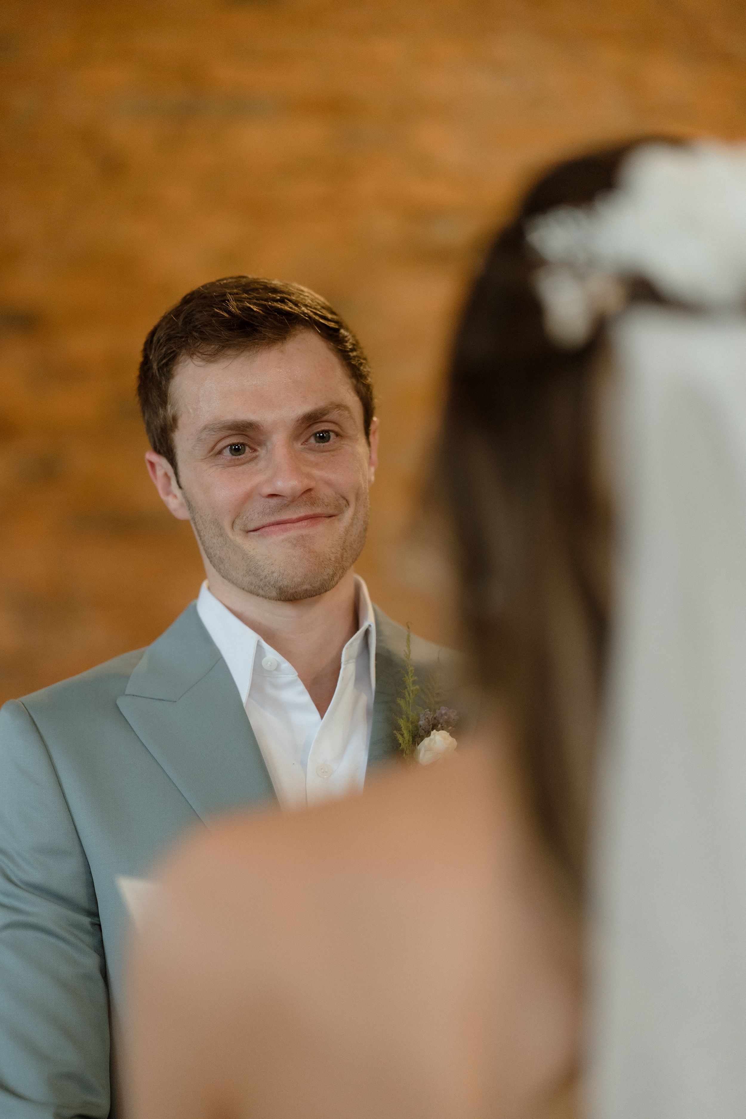 Wedding ceremony candid moment with groom smiling warmly in light gray suit.