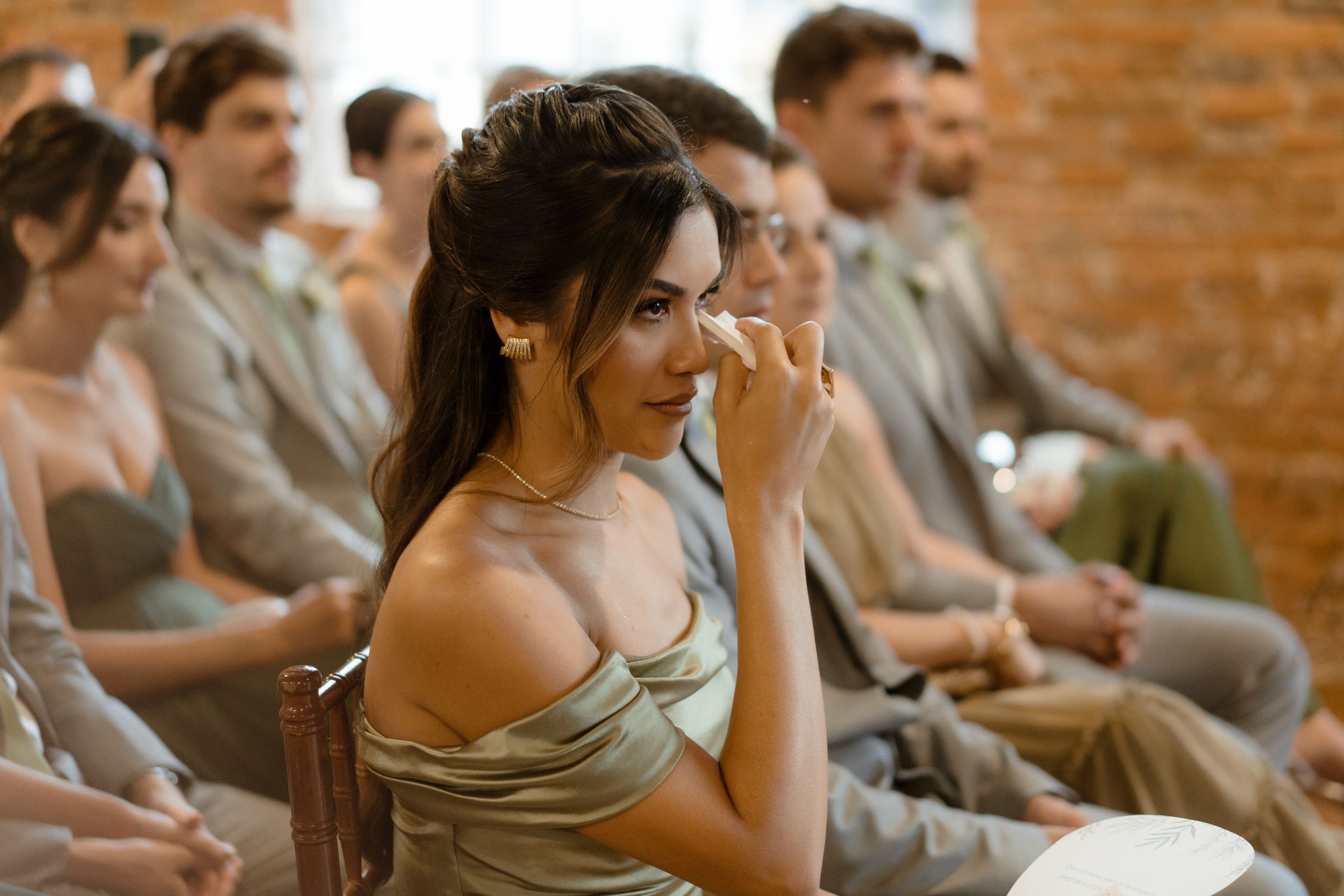 Wedding guest in an off-shoulder dress reacts emotionally during ceremony in rustic brick venue.