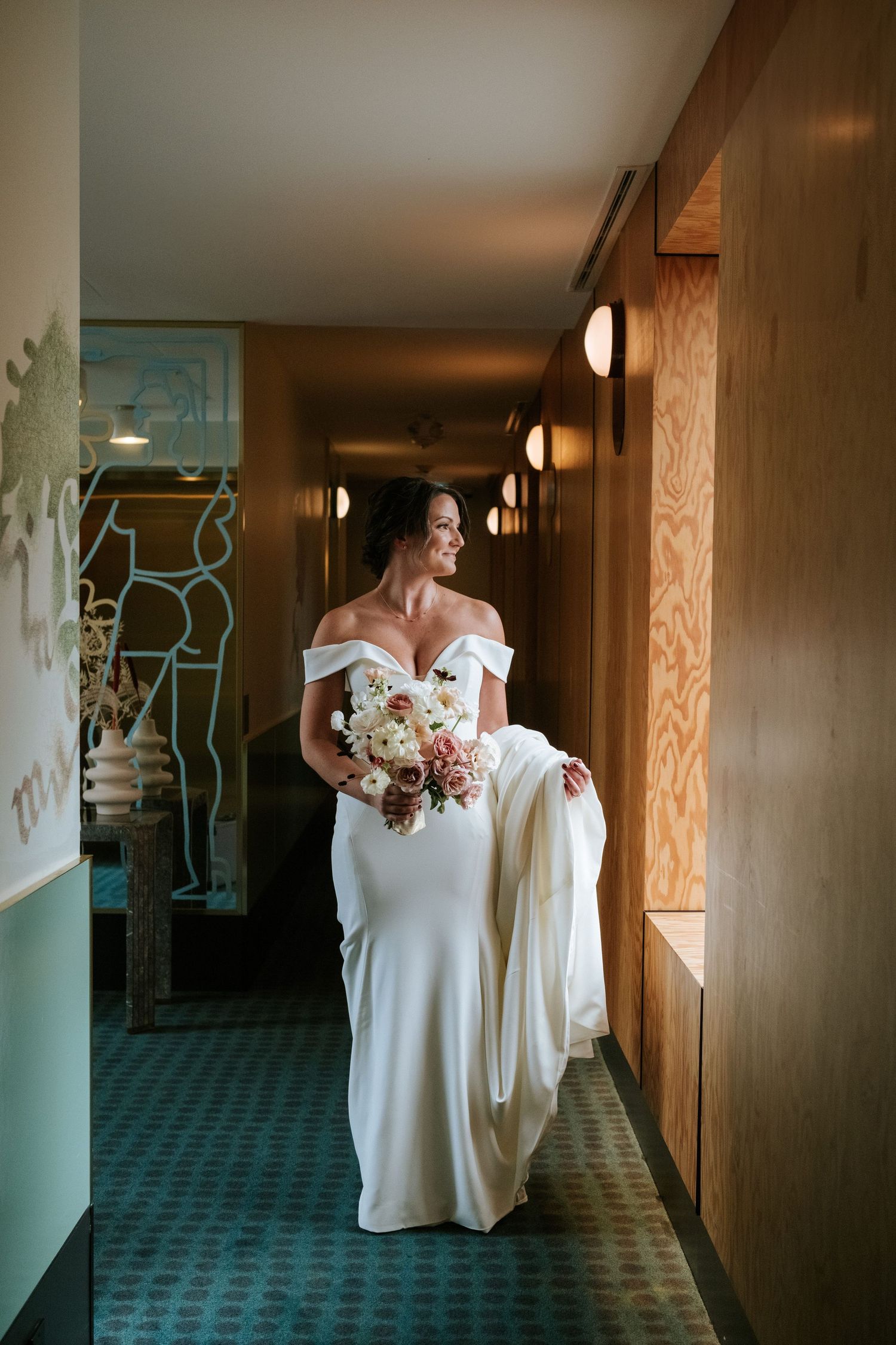 Bride in fitted white gown holding bouquet in modern hotel corridor.