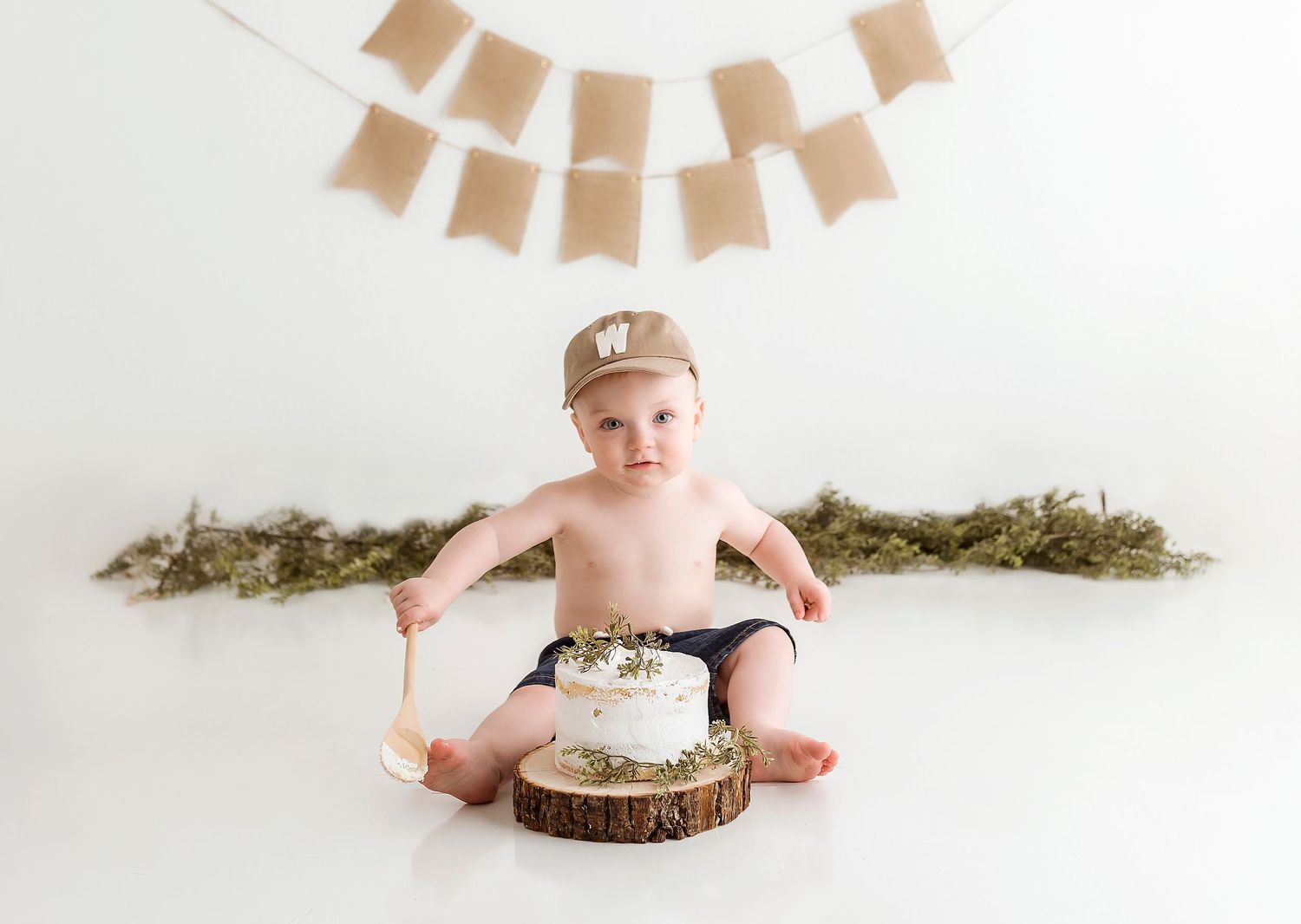 A smash cake setup with rustic wood slice cake stand and greenery garland backdrop for a special celebration.