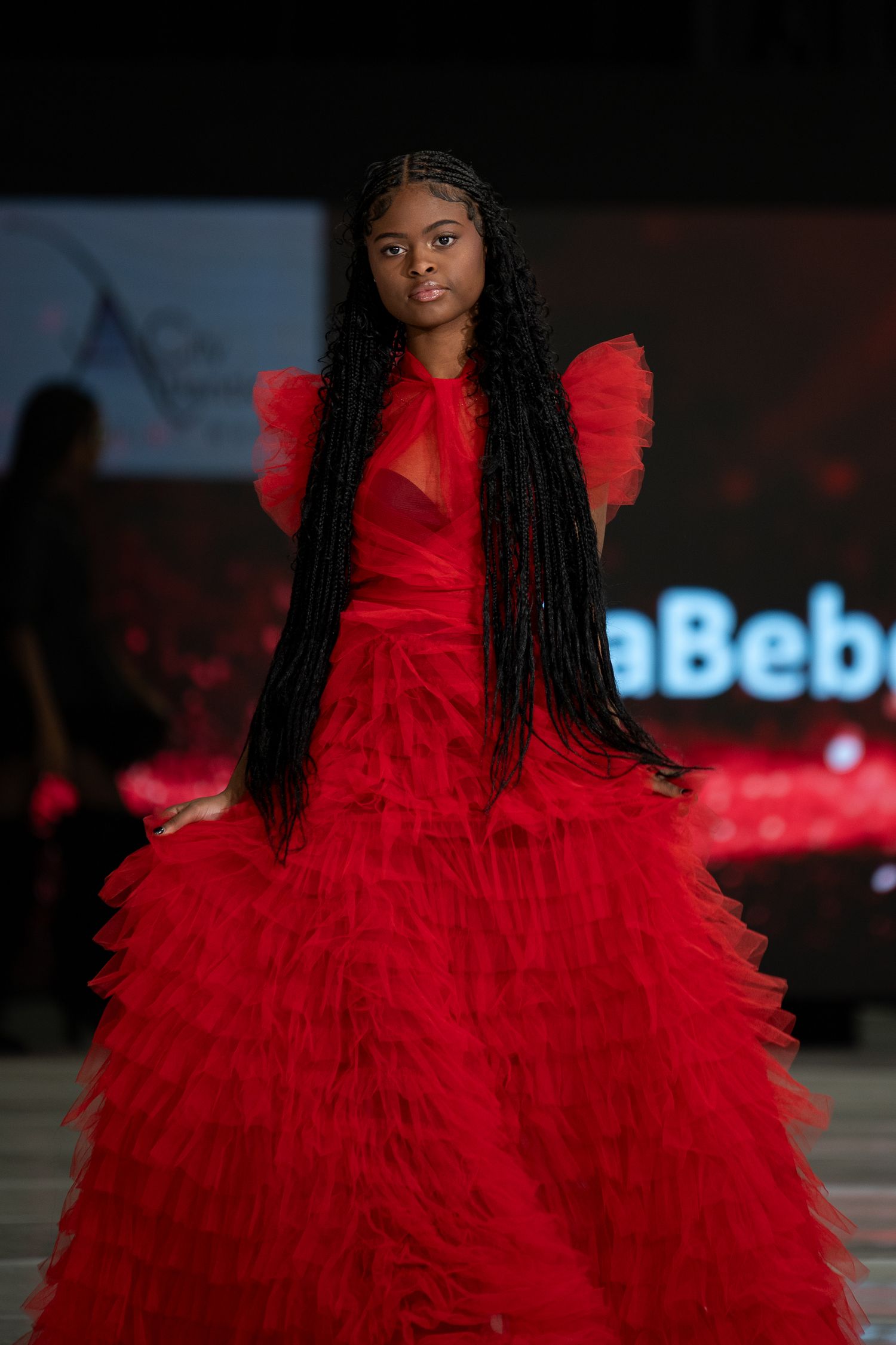 A dramatic red tulle ball gown with ruffled layers and puff sleeves is displayed on a dimly lit fashion runway.
