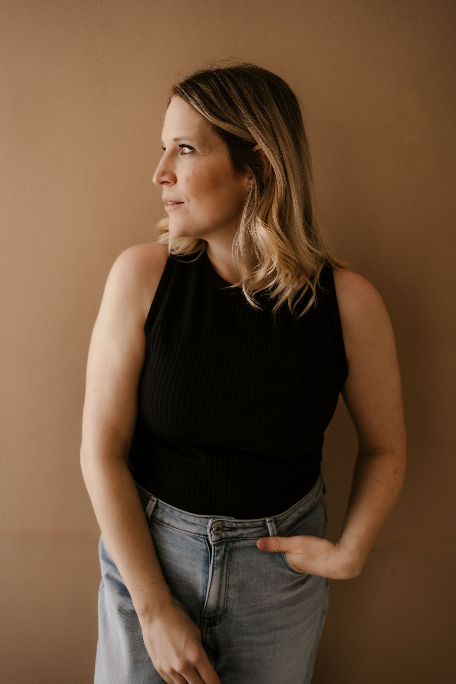 A person in a black sweater and blue jeans sits casually on a white floor in a minimalist setting.