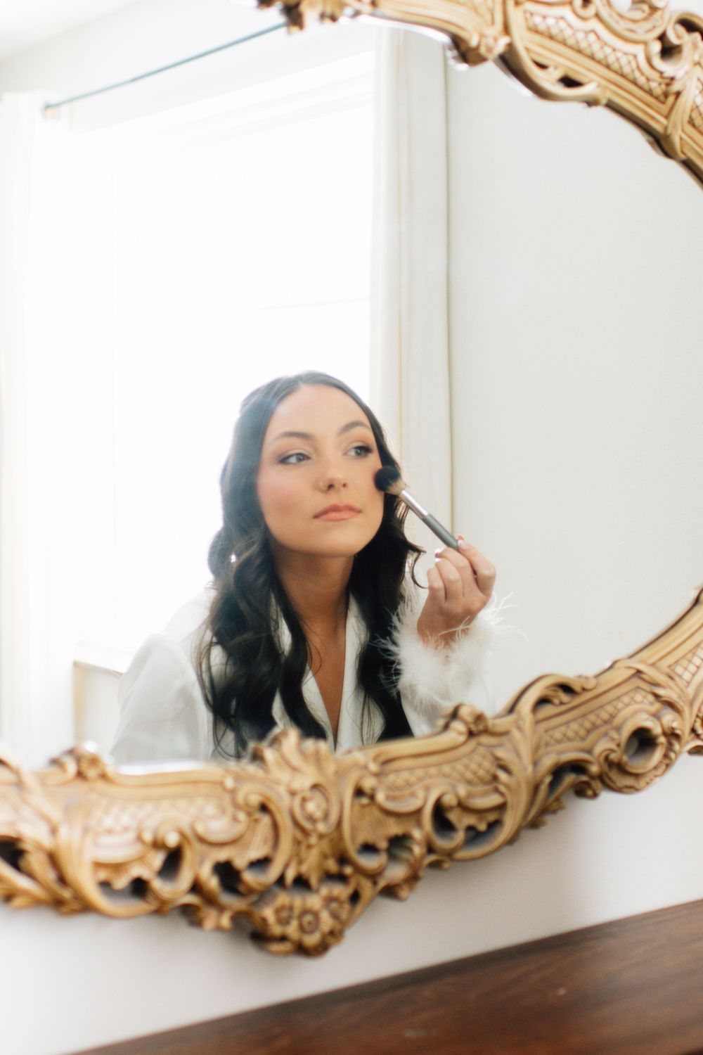 Reflection in ornate gold mirror showing person applying makeup with brush.