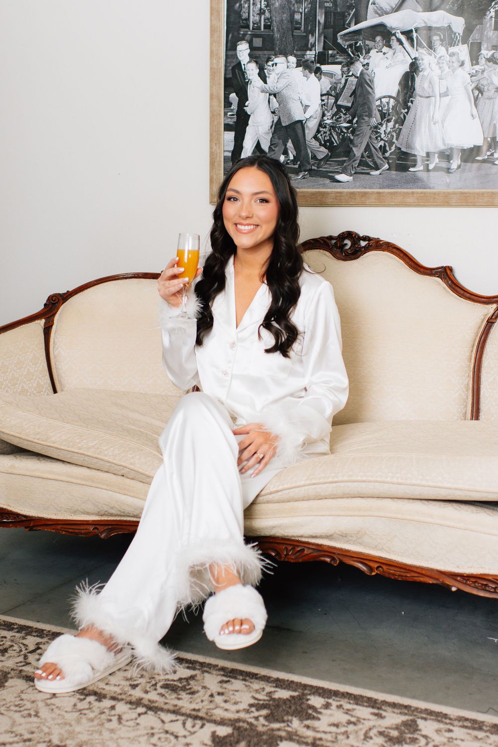 Person in white silk robe relaxing on vintage sofa with champagne glass.