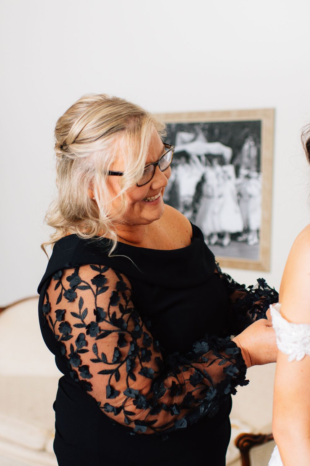 Helper in black lace dress assisting with wedding dress preparations.