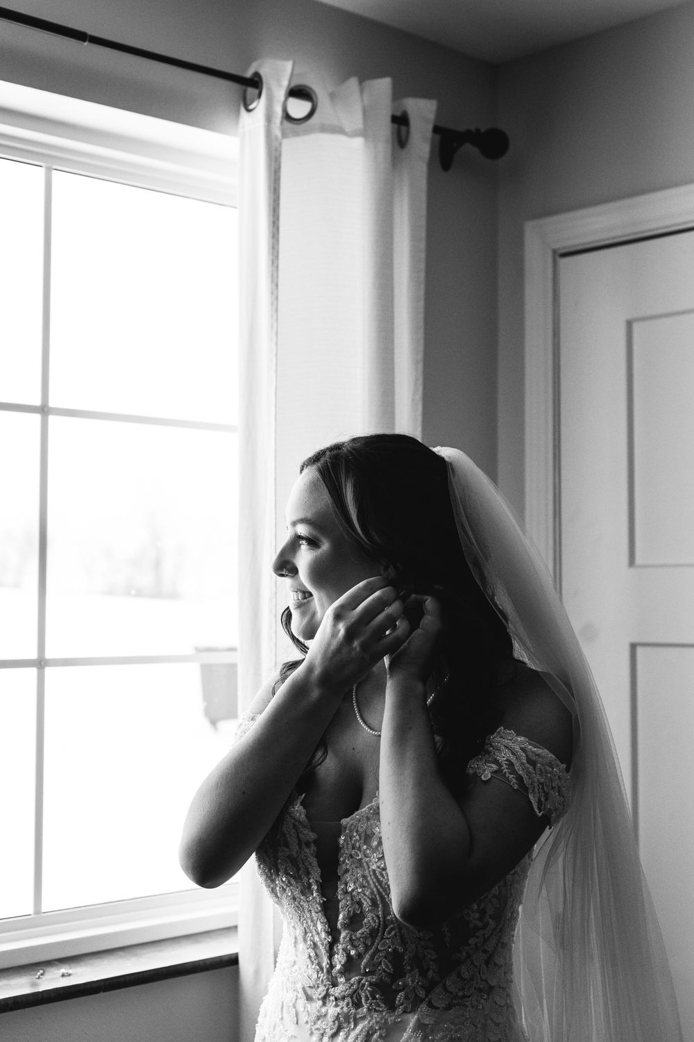 Black and white portrait of bride putting on earrings by window light.