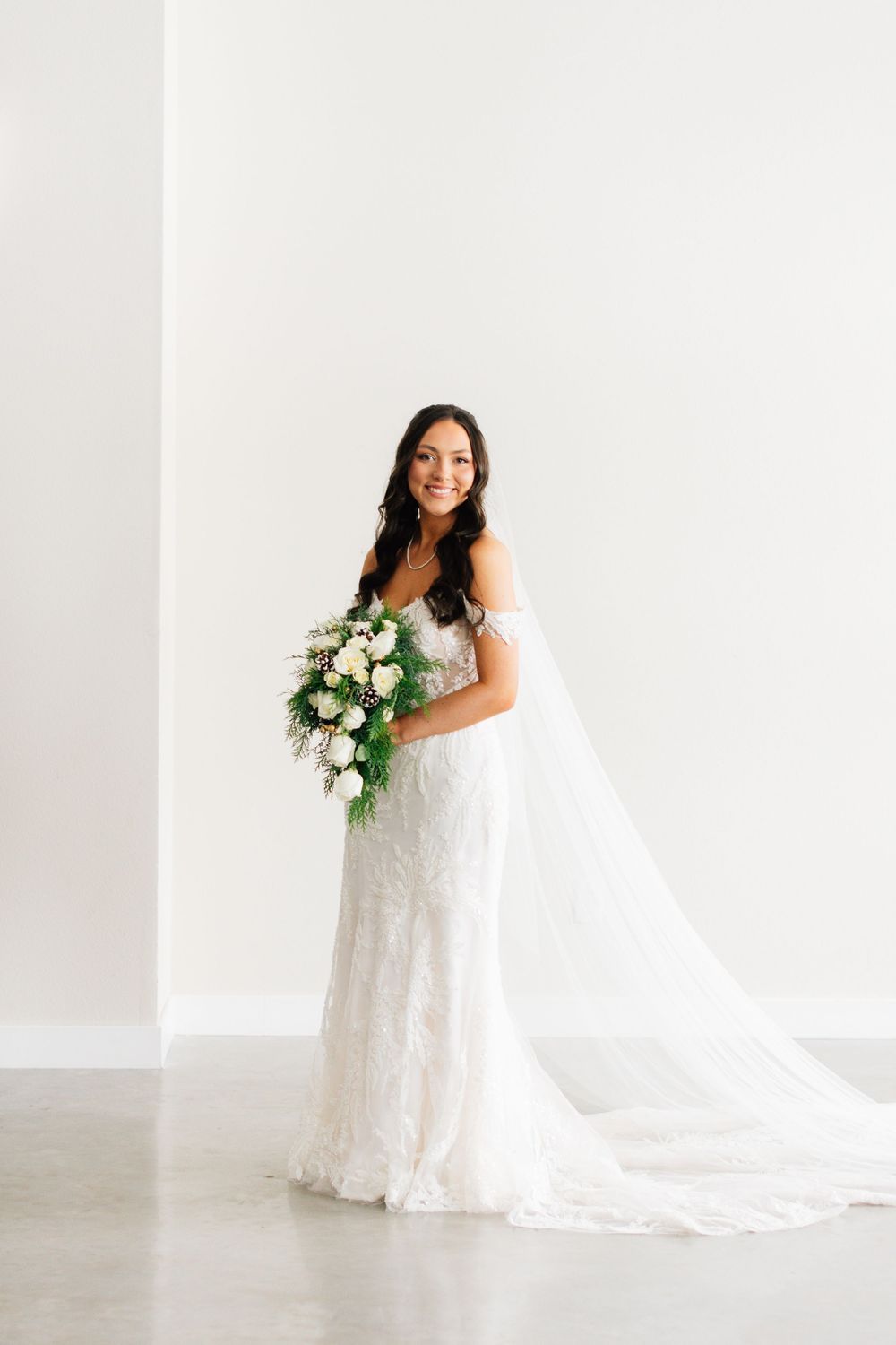 Full length bridal portrait with flowing veil and white floral bouquet against white backdrop.