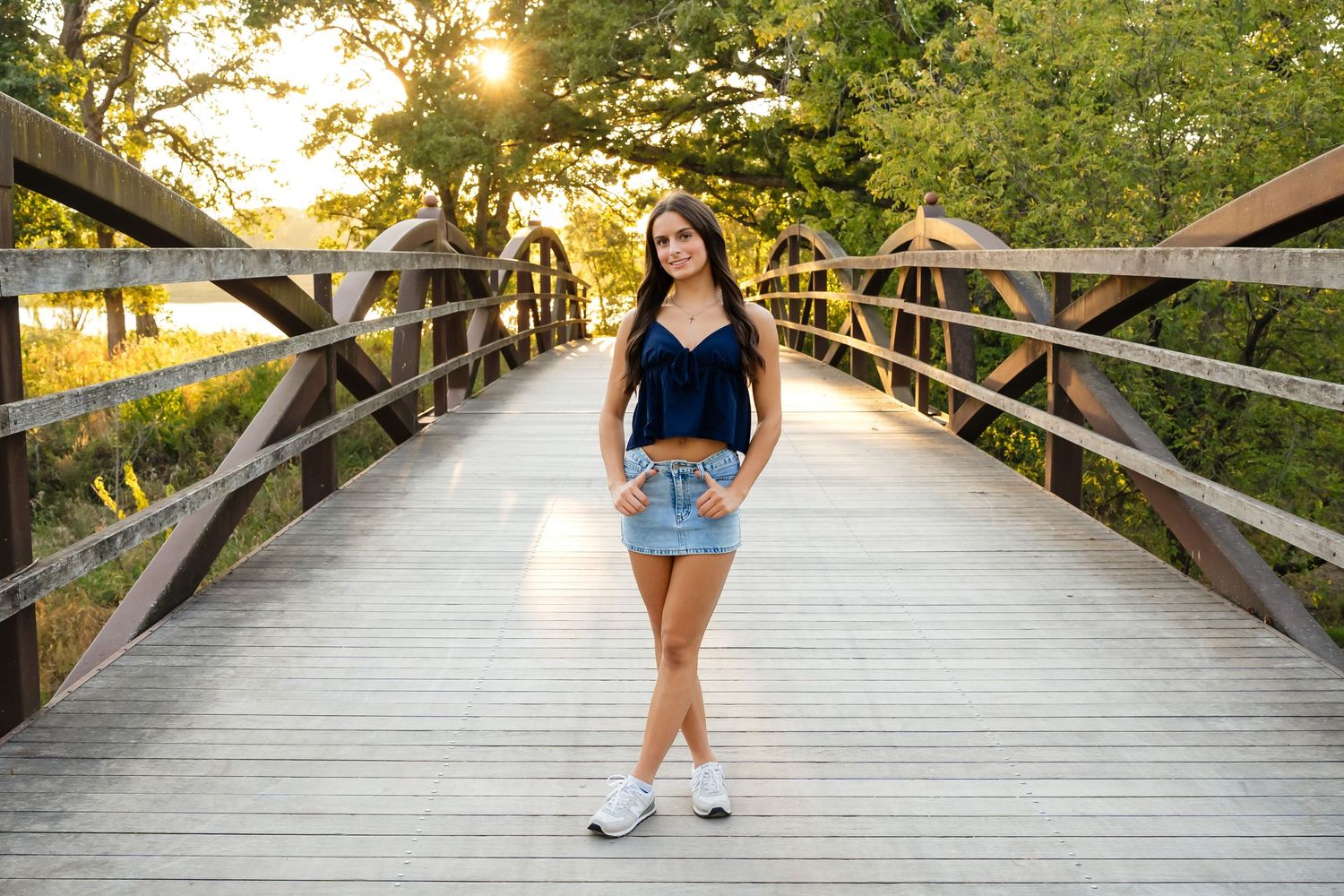 Person walking on a wooden boardwalk path through lush green trees at sunset with golden sunlight filtering through.