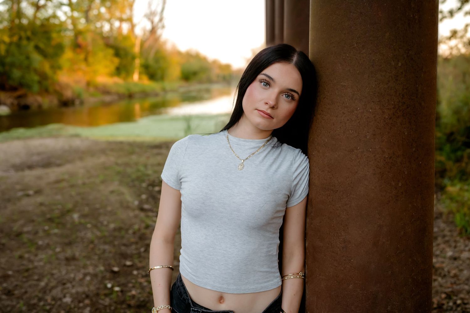 A figure in a light gray crop top poses outdoors during golden hour sunset in a natural setting.