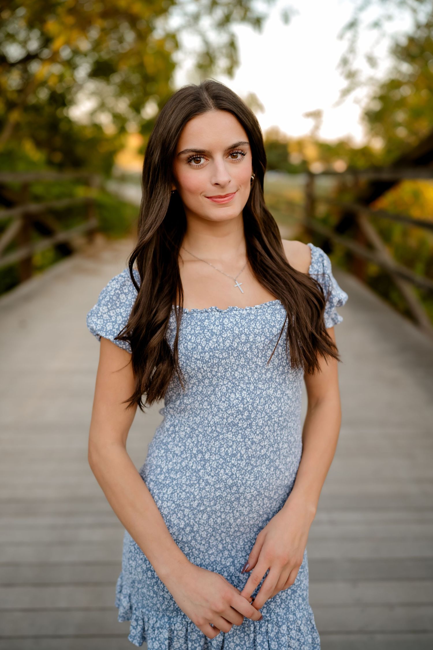A woman in a blue floral dress poses outdoors on a wooden boardwalk during golden hour.