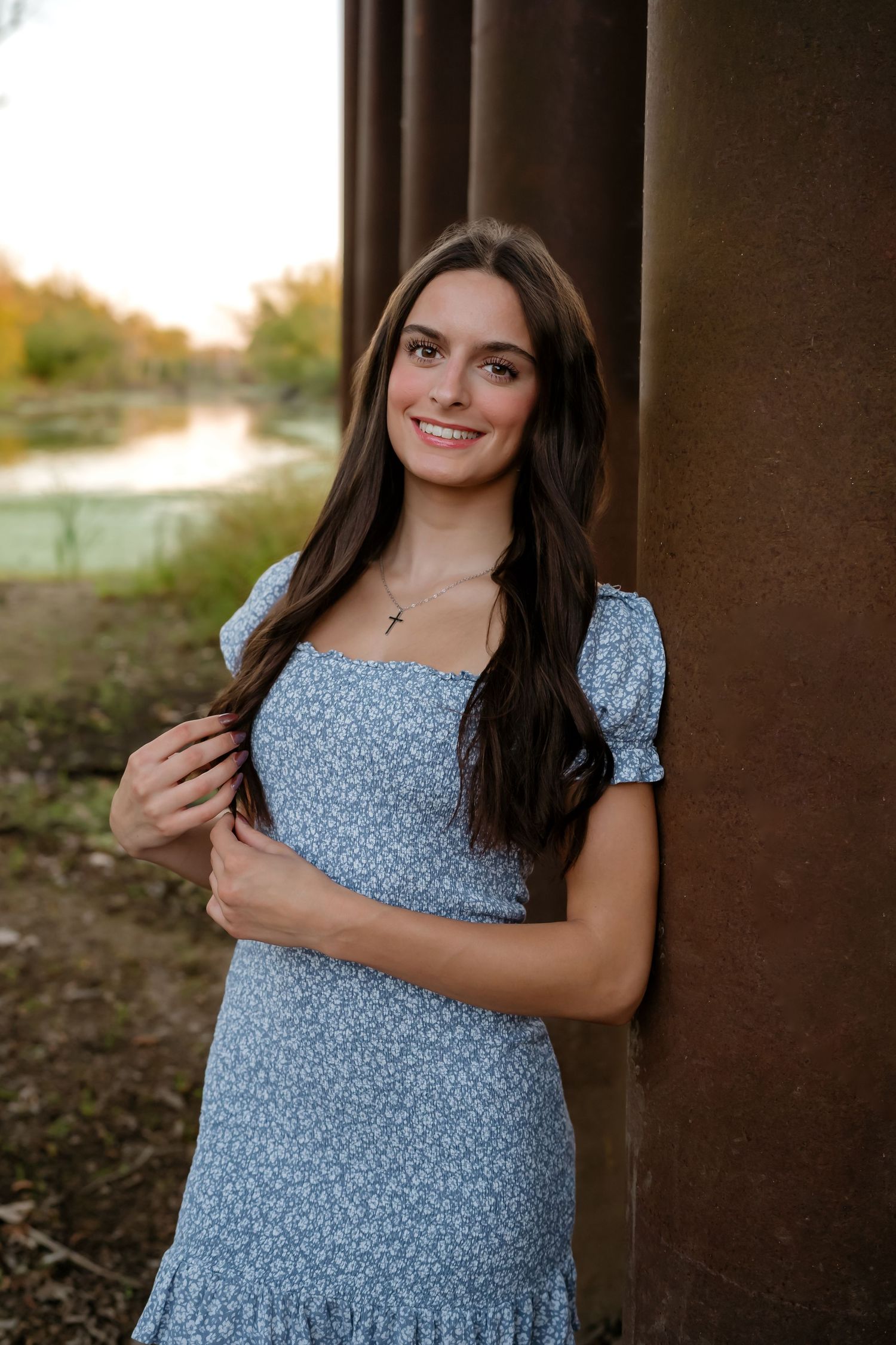 A person in a light blue floral dress poses outdoors against a rustic backdrop during golden hour.