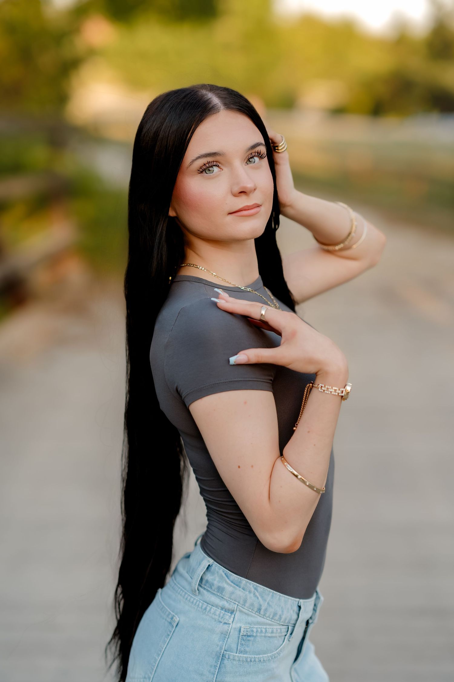 Young woman with long black hair poses outdoors in casual attire against a blurred natural background.