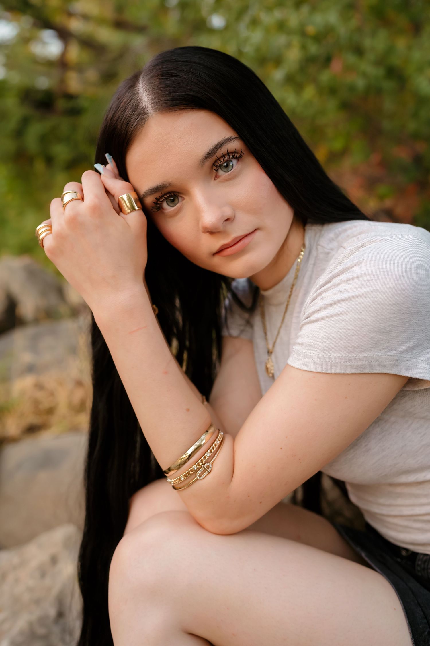 A person with long dark hair poses outdoors against a natural backdrop in soft lighting.