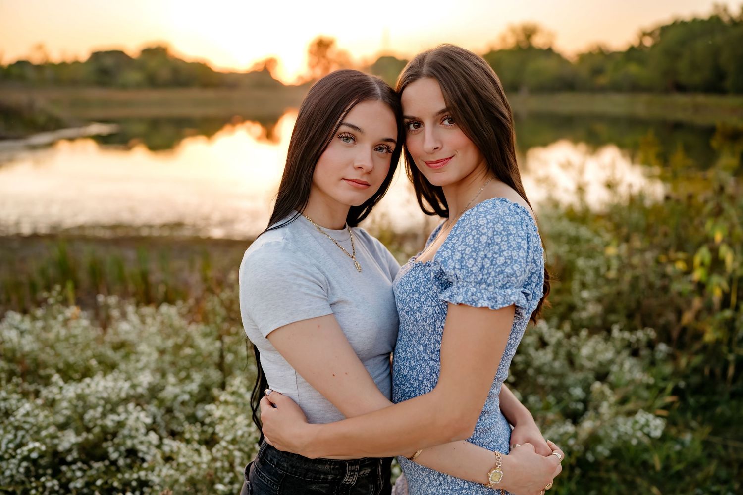 Two people embrace during golden hour sunset in a field of wildflowers beside a tranquil lake.