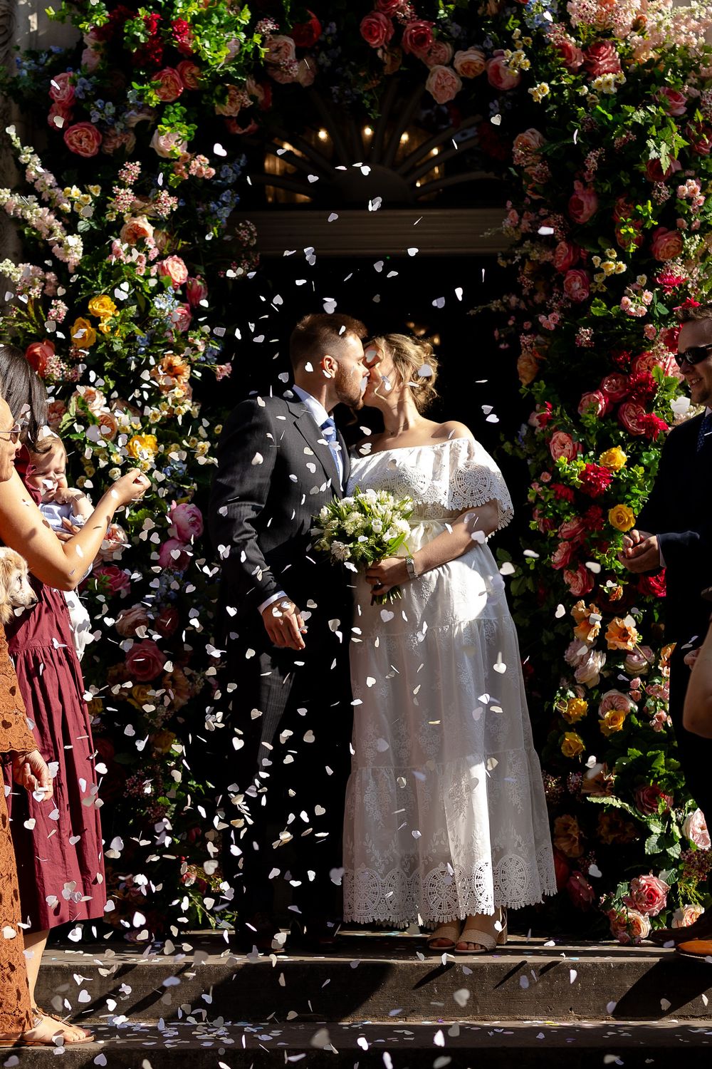 A romantic wedding ceremony under a lush floral arch with white petals falling during a joyful celebration.