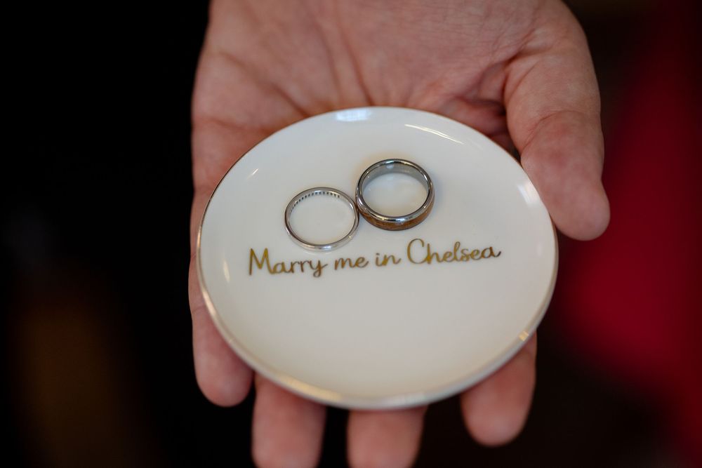 Two wedding rings rest on a white ceramic dish with 'Marry me in Chelsea' written in gold script.