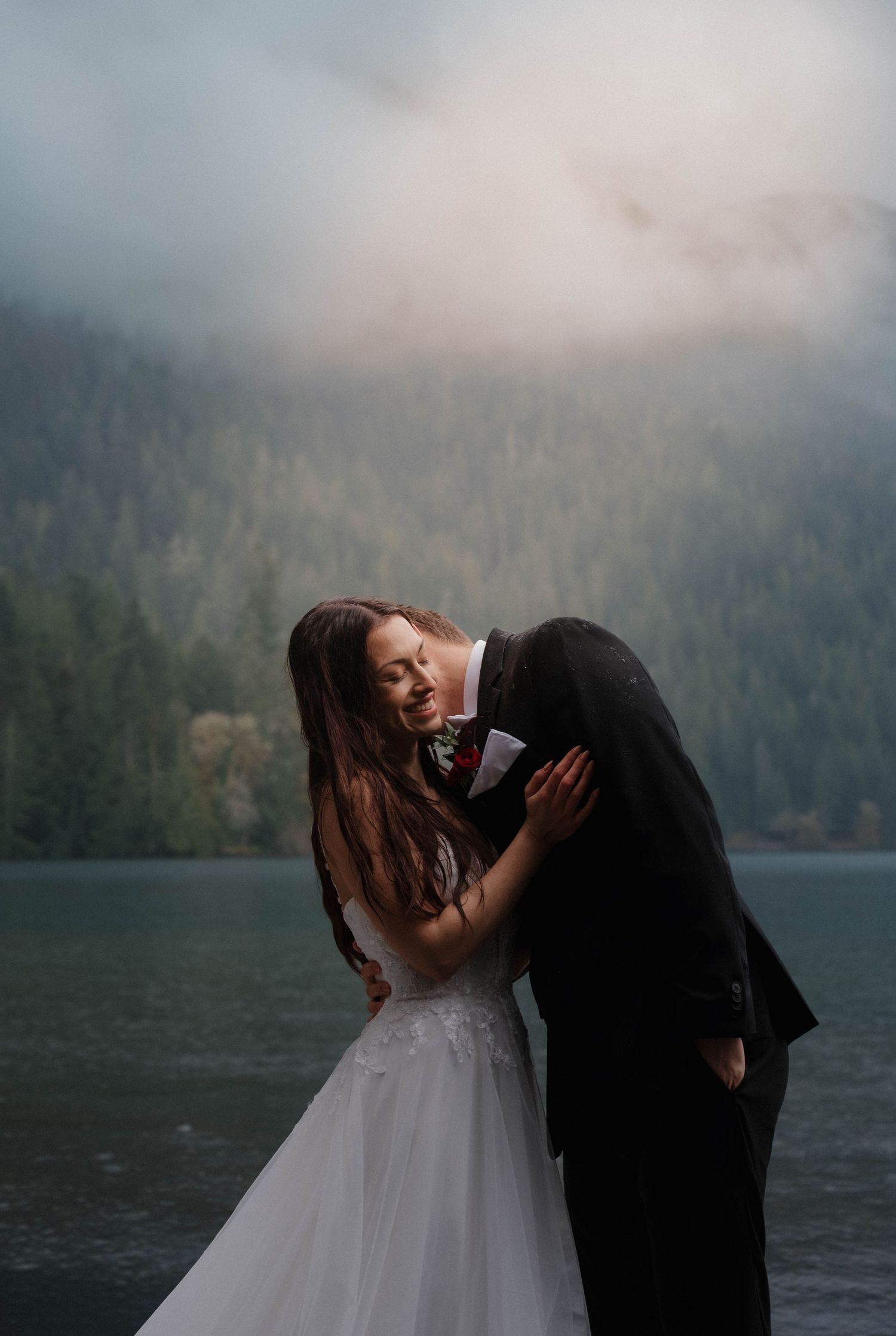 Bride and groom embrace romantically beside a misty mountain lake during a moody outdoor elopement.