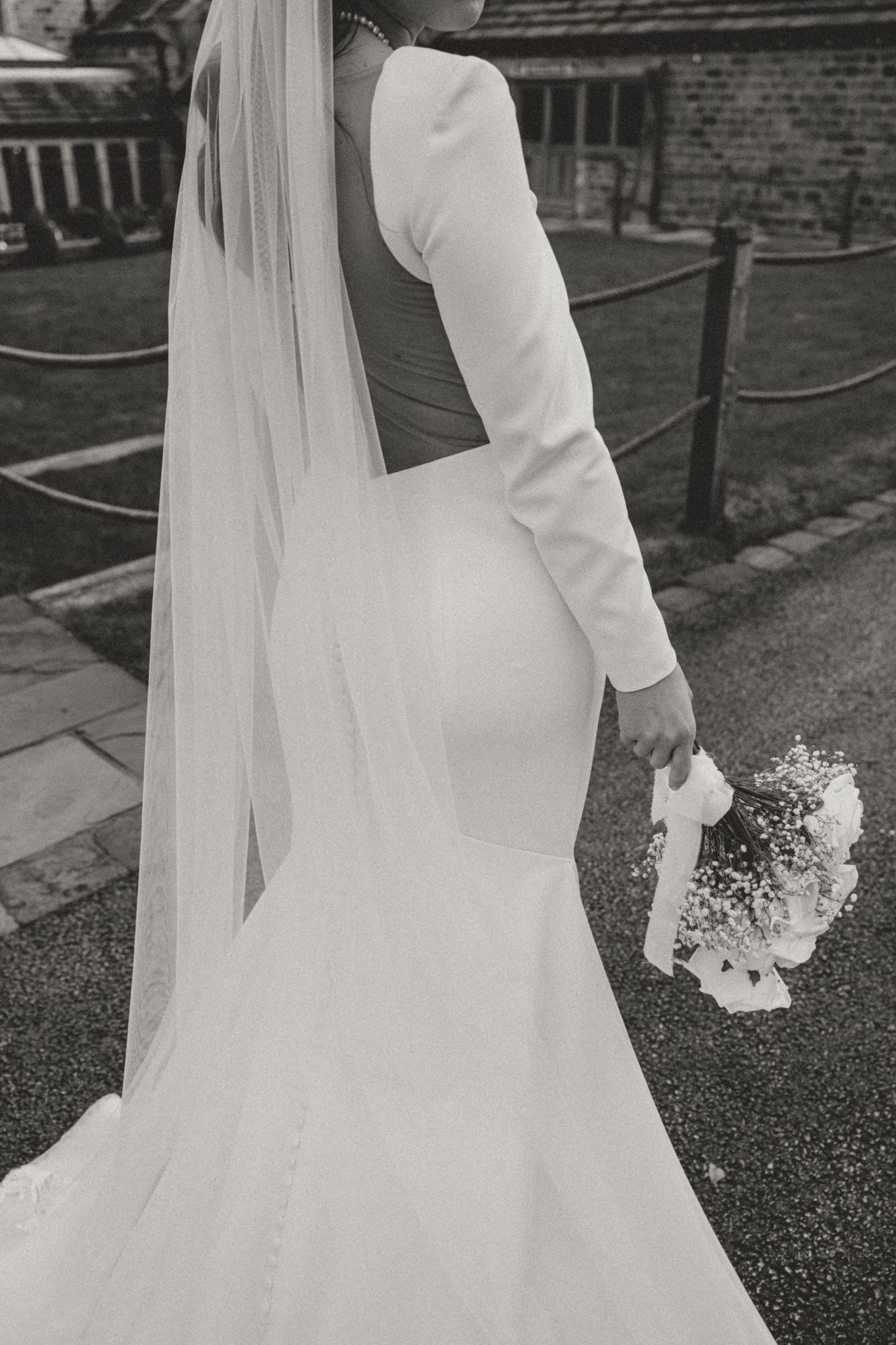 Black and white image of a bride in a long white wedding dress and veil holding a bouquet while walking.