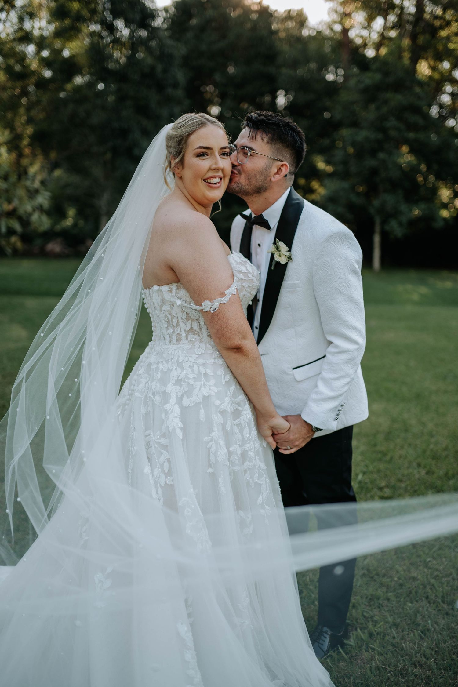 A romantic wedding couple embraces outdoors on their special day as sunlight filters through trees in the background.