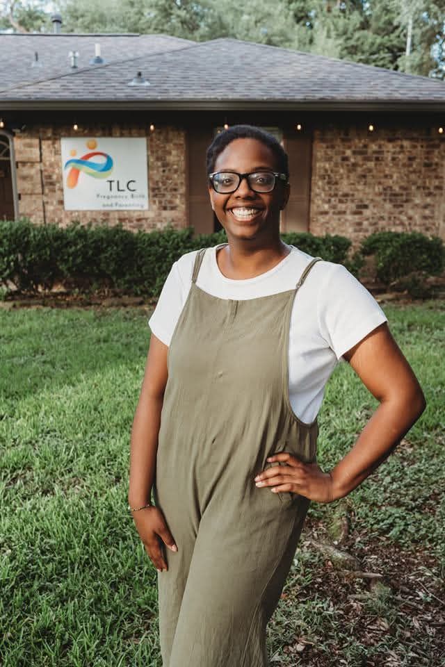 A person with brown skin and black hair in a casual updo, who wears glasses, is smiling and standing with their hand on their hip.