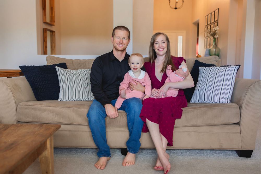 A family sits together on a beige couch in a living room with two small babies in pink outfits.