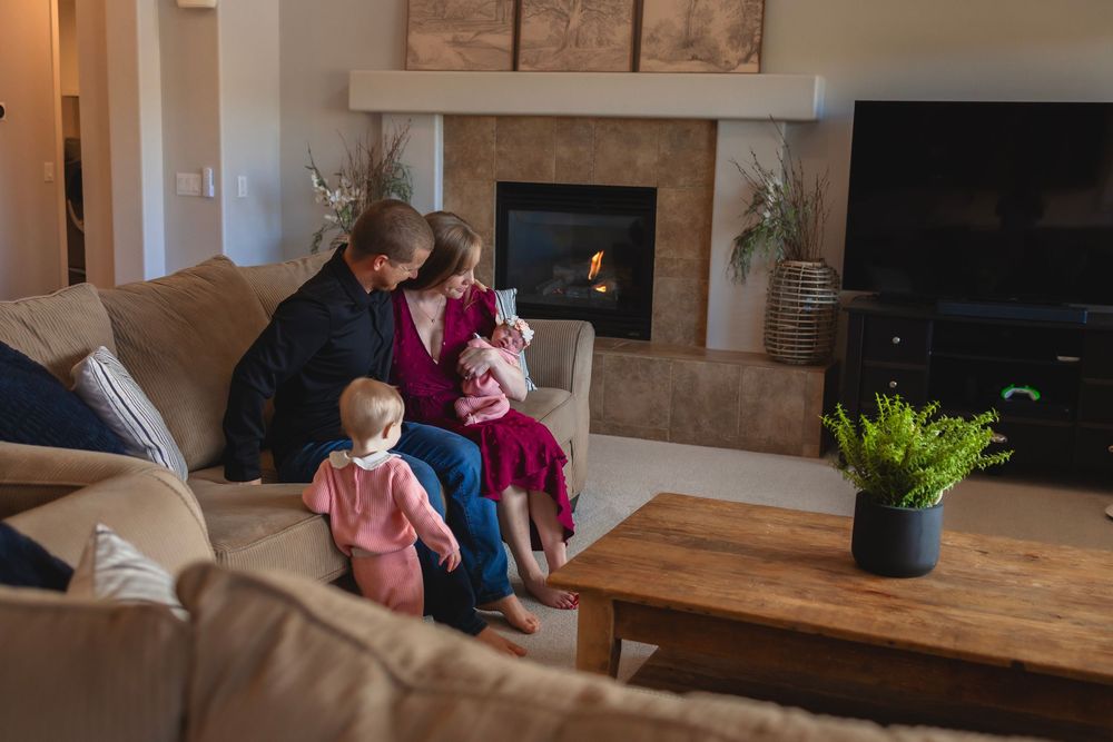 Family of 4 with newborn baby relaxes together on a comfortable gray sectional sofa in a modern living room with natural light.
