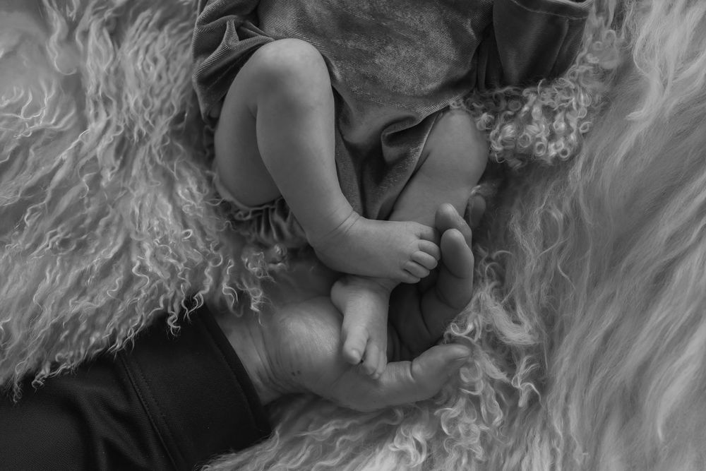 A black and white photo showing tiny baby feet nestled in soft fabric, fathers hand holding her little feet