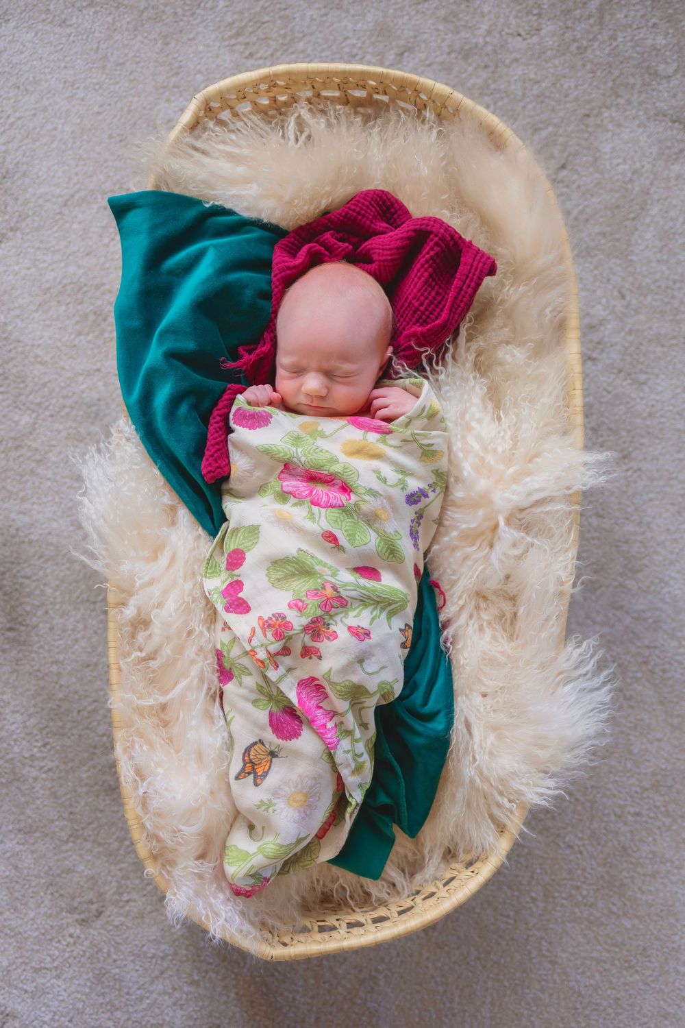 A newborn baby sleeps peacefully in a cream-colored basket with teal and pink fabric accents.