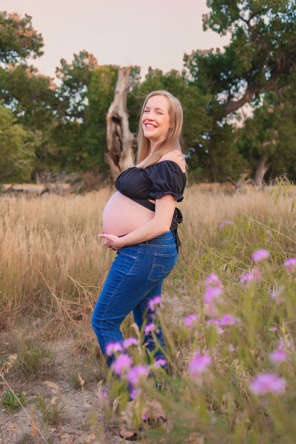 Expectant mother in blue jeans and black top poses in a field with purple wildflowers during golden hour.