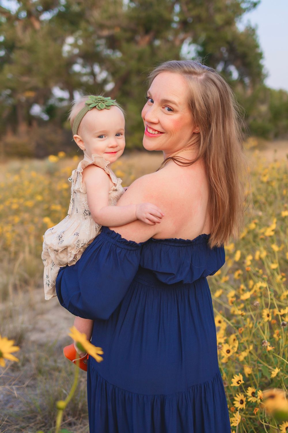 A woman in a navy off-shoulder dress holds a baby in a floral dress amidst yellow wildflowers on a sunny day.