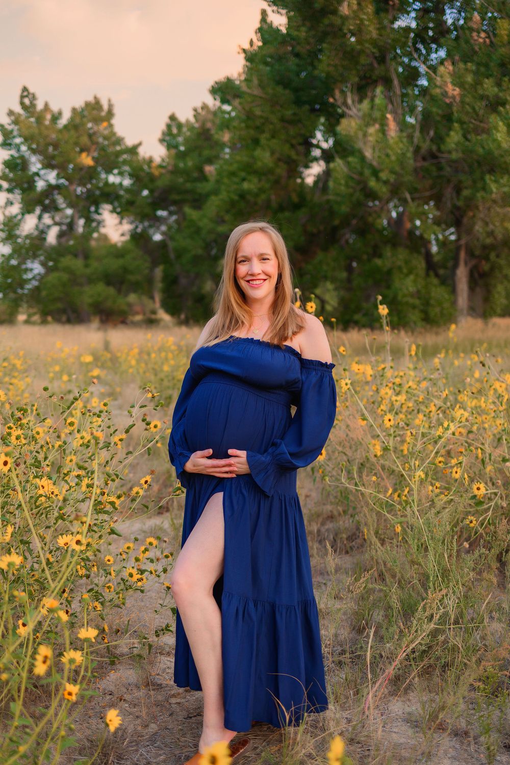 A maternity photo in a field of yellow wildflowers at sunset shows an off-shoulder navy blue dress against a natural backdrop.