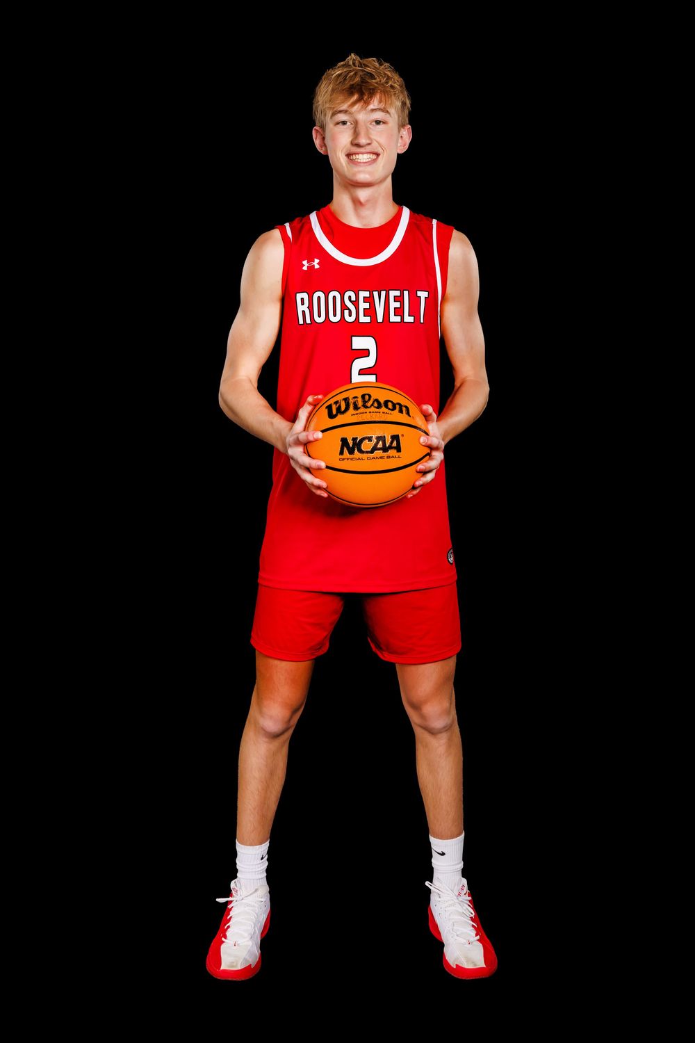 Basketball player in red Roosevelt uniform crouches with ball against black background in sports portrait pose.