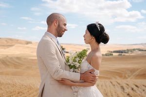 Wedding couple embraces in a sun-drenched wheat field with dramatic rolling hills and blue sky backdrop.